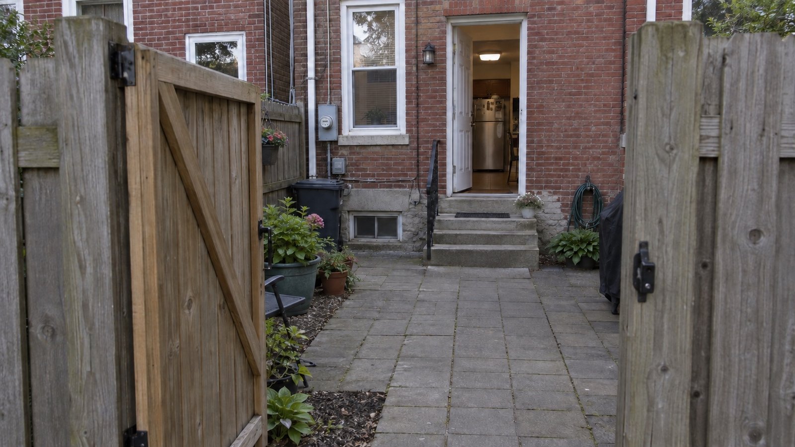 An open rear gate leading across a paved yard to steps and an open kitchen door.