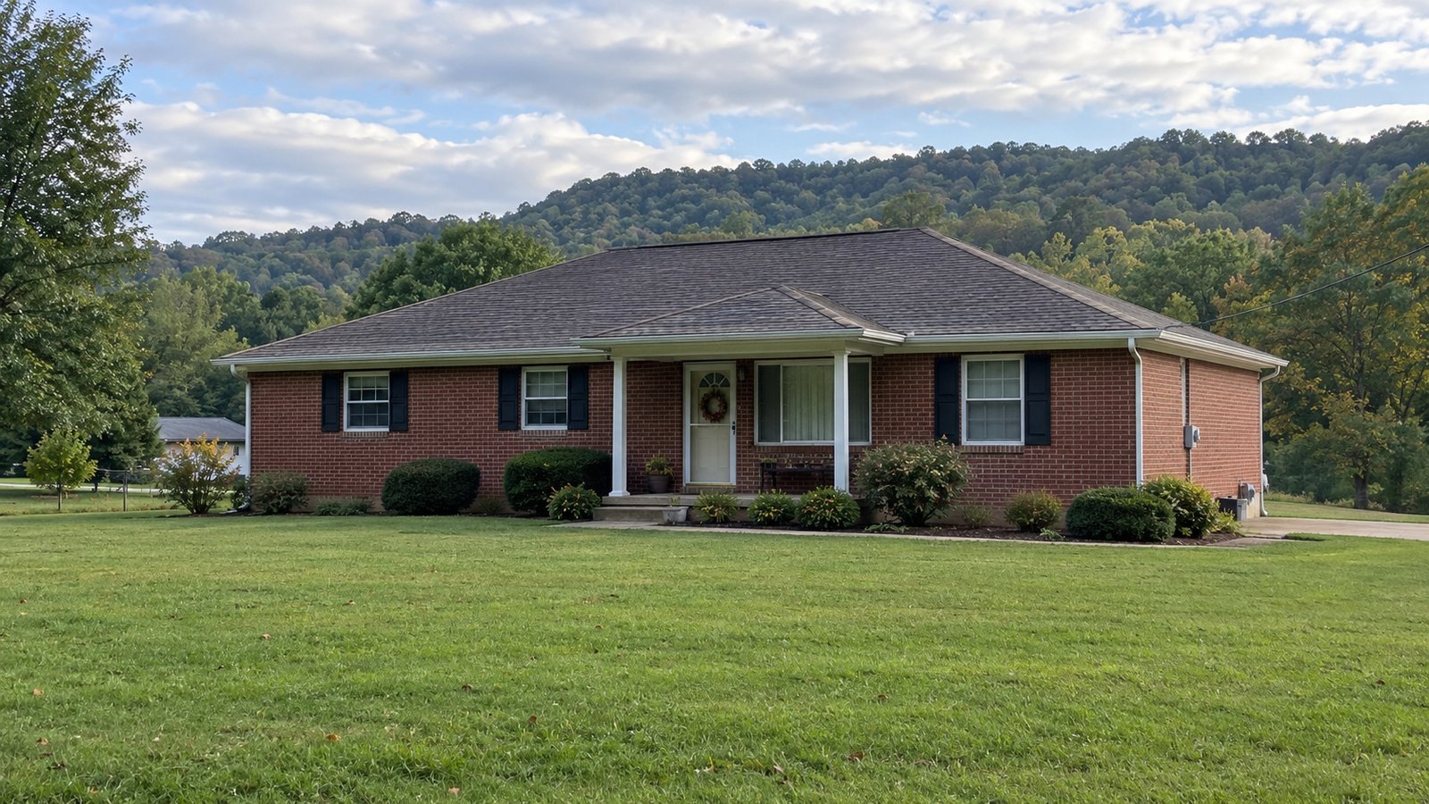 A modest brick home near wooded hills A complete one-story brick house with a covered entrance and a broad lawn