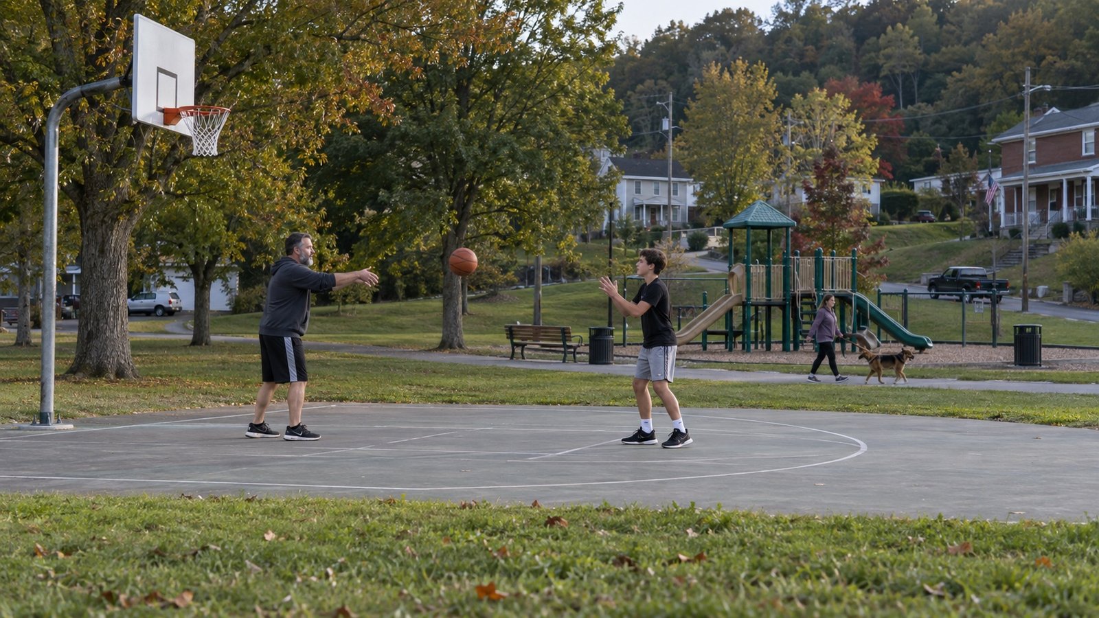 A parent and teenager passing a basketball on a park court, with a path and playground beyond.