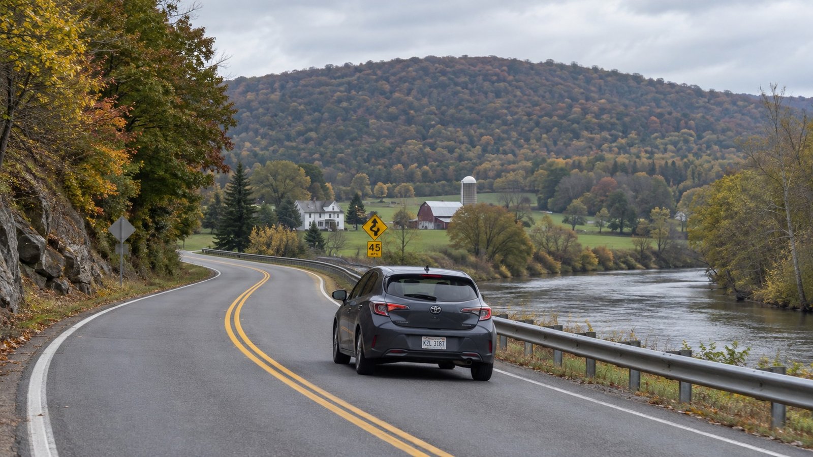 A compact car traveling in the right lane of a curving regional road beside a river, with a curve advisory sign.