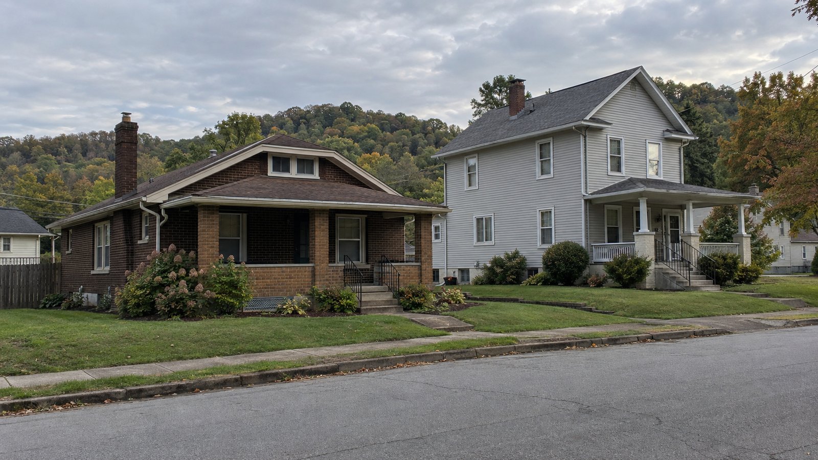 A complete brick bungalow and gray two-story home beside a residential street with wooded hills behind.