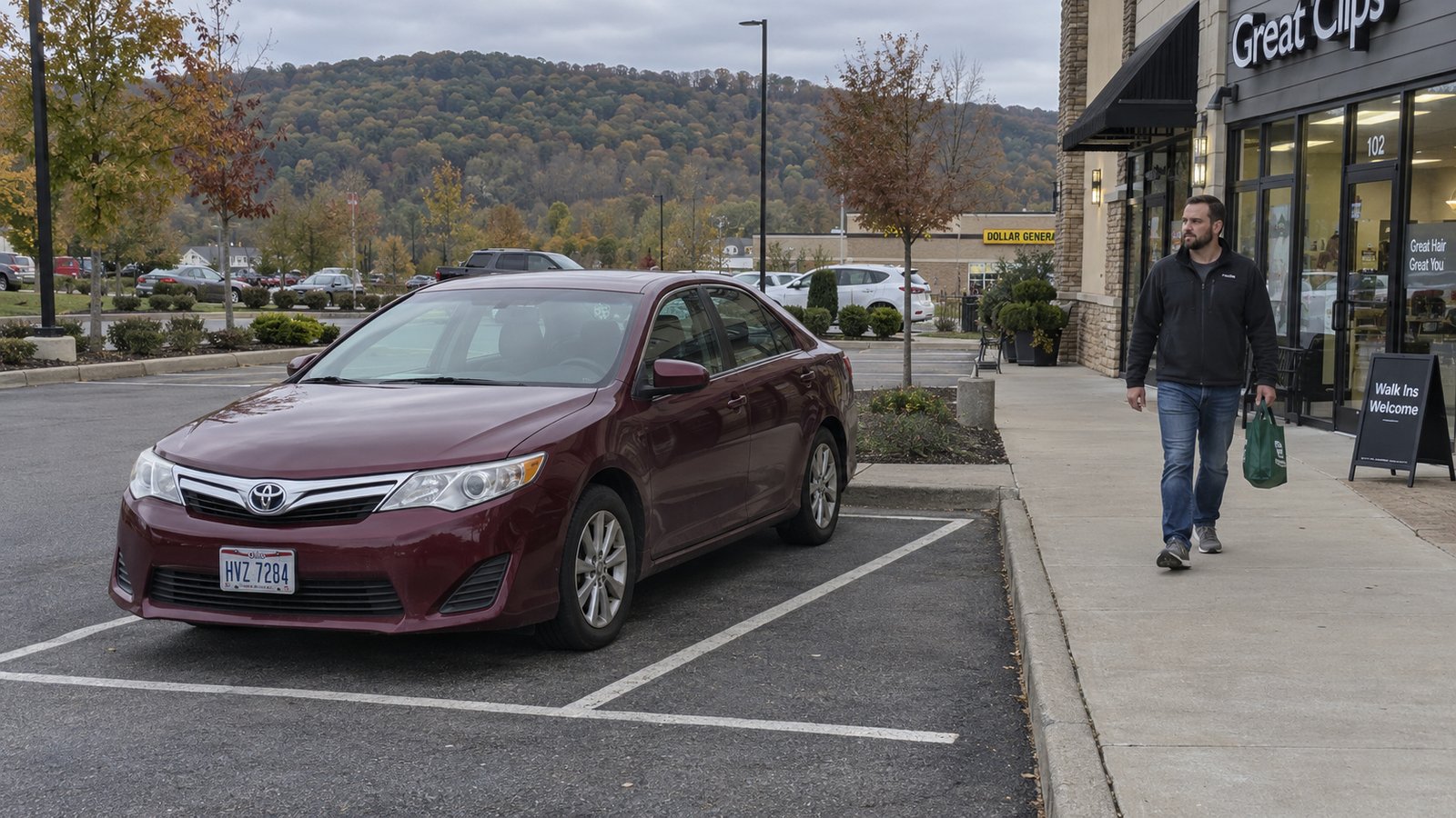 A man with a reusable bag walking along a raised sidewalk beside a complete red sedan in a marked parking bay.