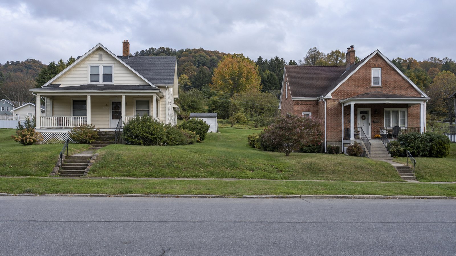 Two complete modest homes, one cream-sided and one brick, beside a residential street and grassy rise.