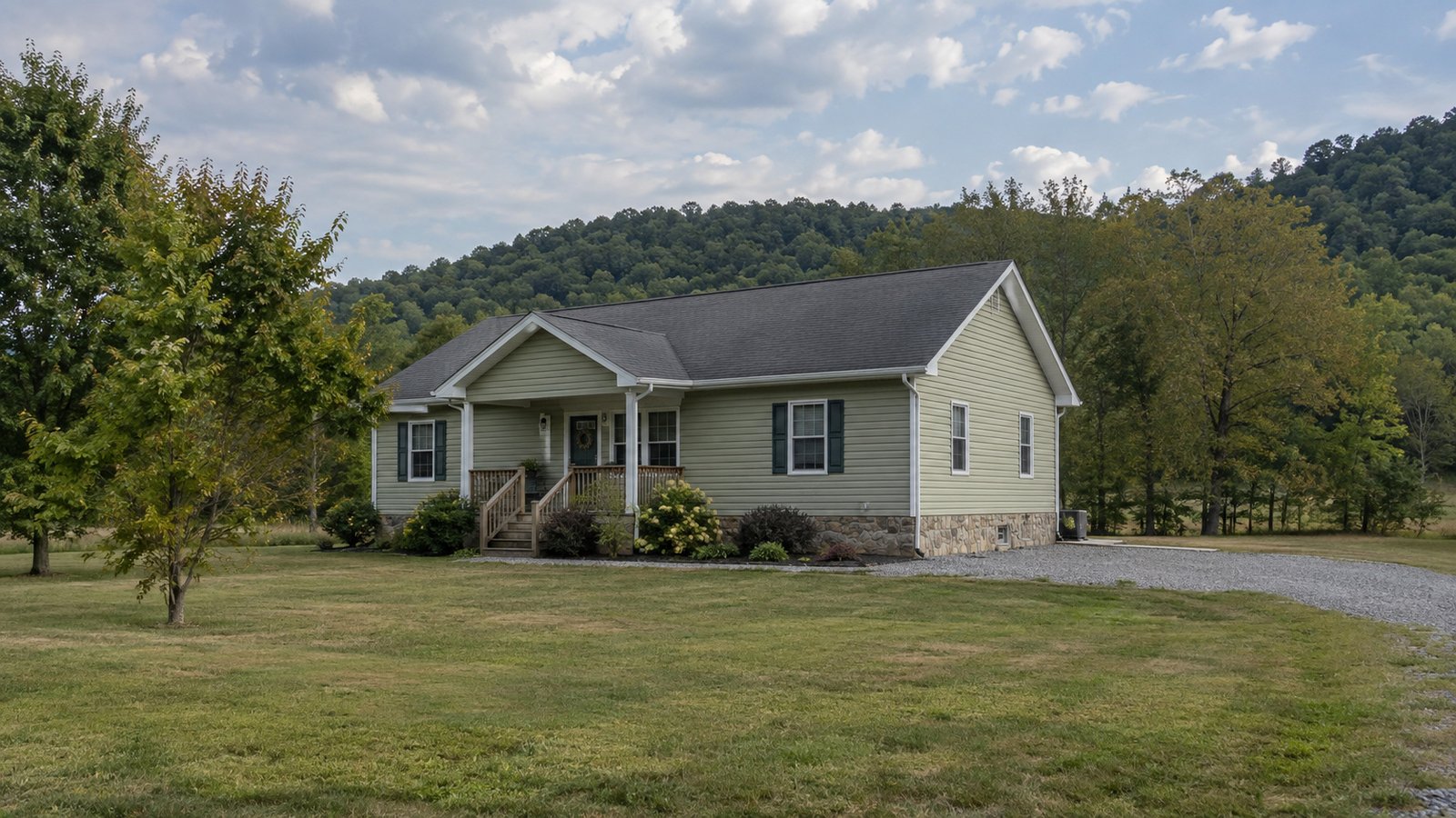 A whole pale sage single-story house with a stone foundation, front porch and gravel driveway below wooded hills.
