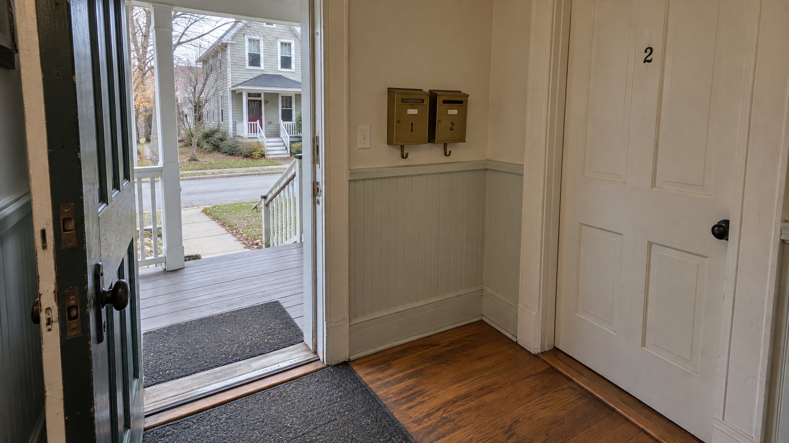Shared vestibule with two mailboxes, a closed apartment door and an open outer door leading to the porch and street