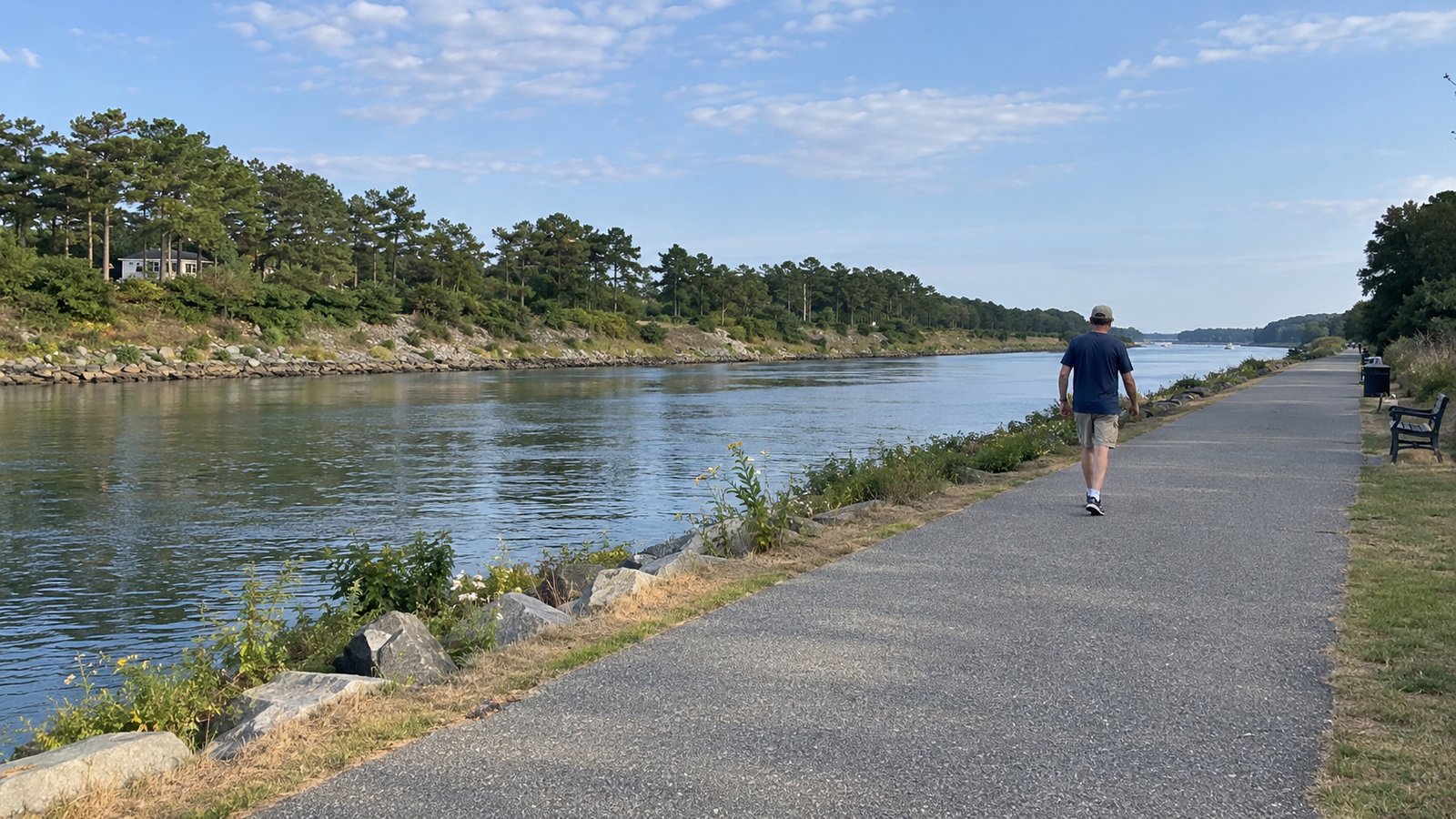 A man walking on a paved waterside path beside calm water and a wooded bank.