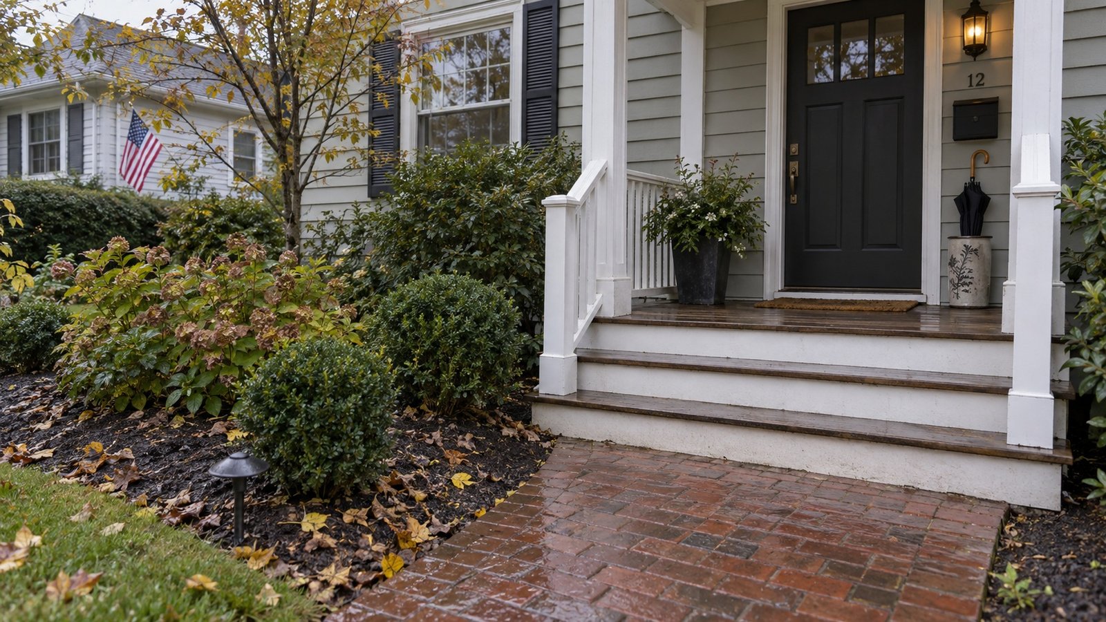 Damp brick path leading to wooden porch steps, with fallen leaves in the garden and an umbrella beside the door