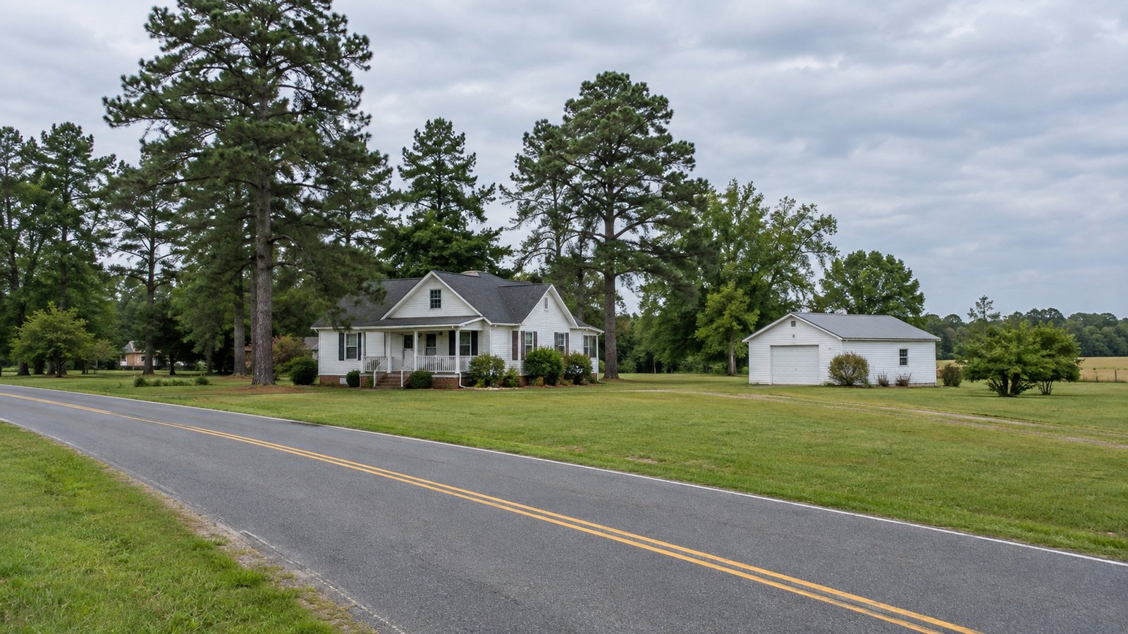 A white clapboard home and detached garage stand behind a grassy verge beside a quiet paved road