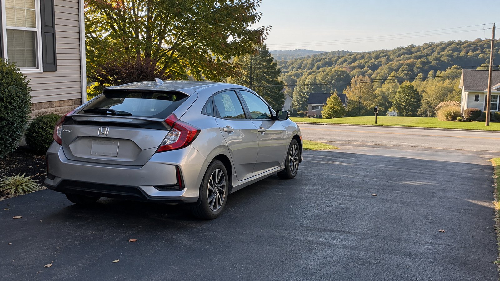 Silver hatchback parked on a home driveway facing a quiet road with wooded hills beyond