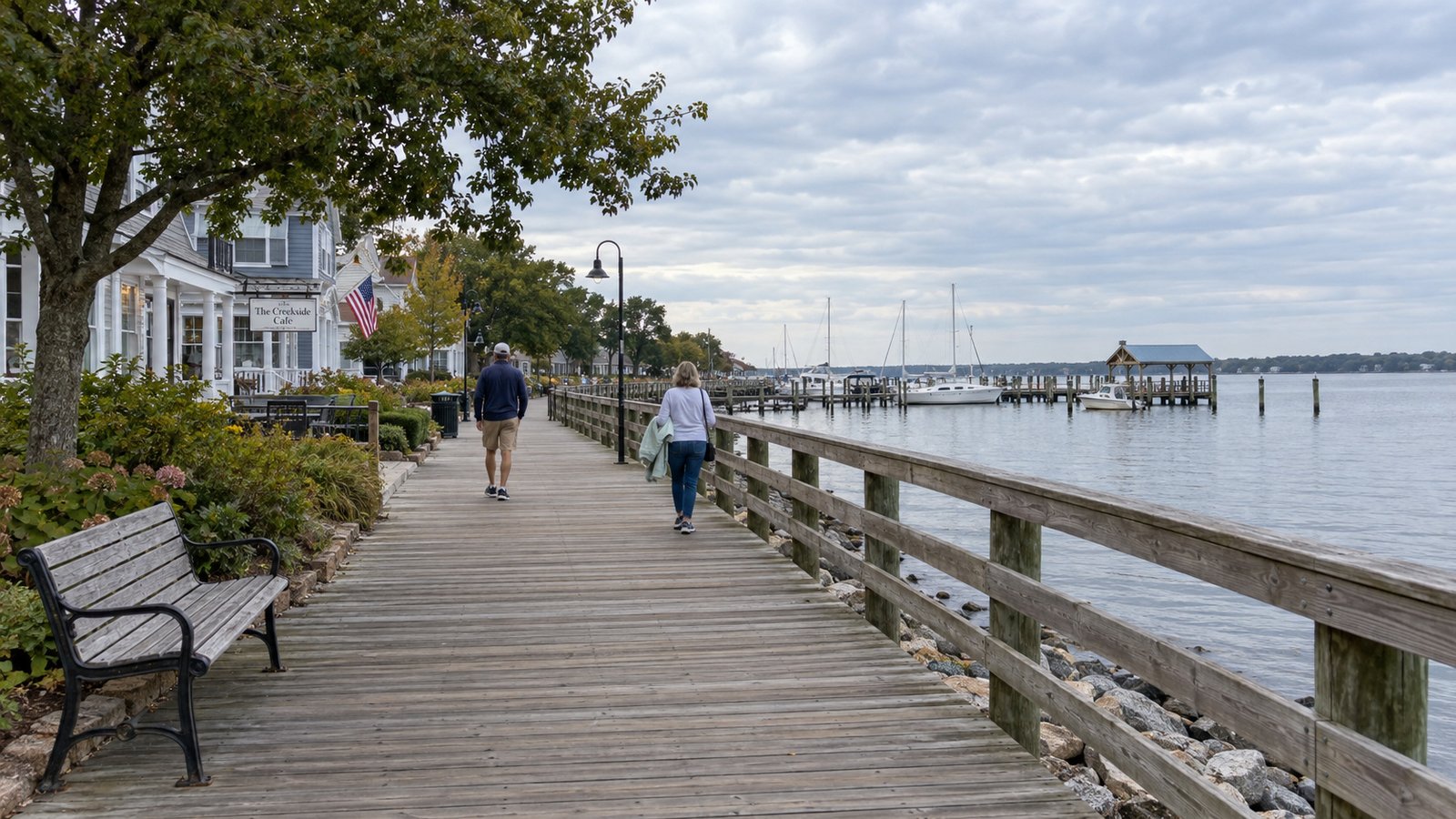 Two pedestrians on a waterfront boardwalk beside homes, a café and moored boats