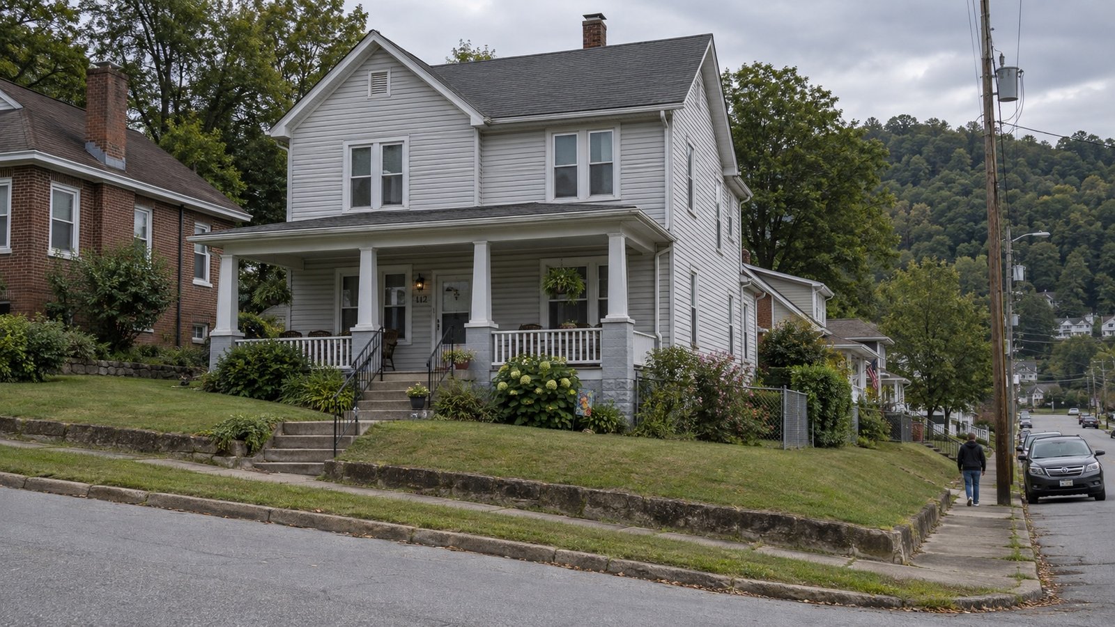 Gray clapboard house with a covered porch, front steps and a wooded hillside behind neighboring homes
