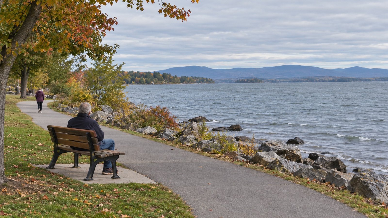 Plan an ordinary outdoor visit A complete bench beside a lakeshore walking path, with one seated adult and another walking farther along the path.