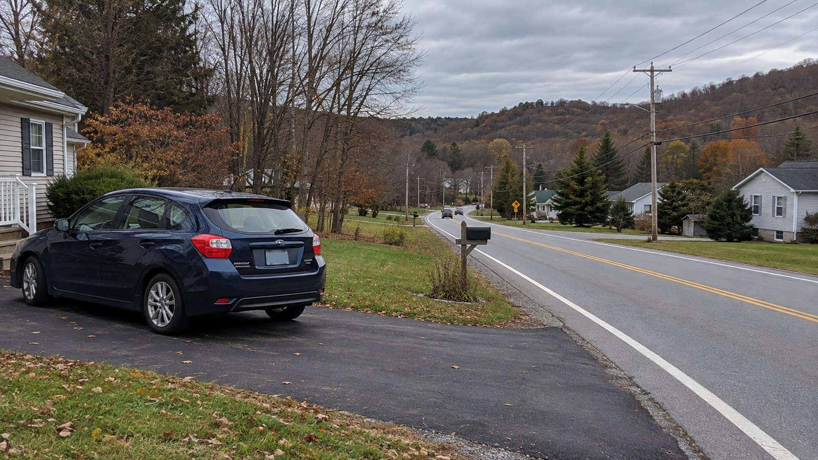 A driveway connected to a village road Dark blue hatchback parked fully on a paved residential driveway beside a two-lane road and low wooded hills