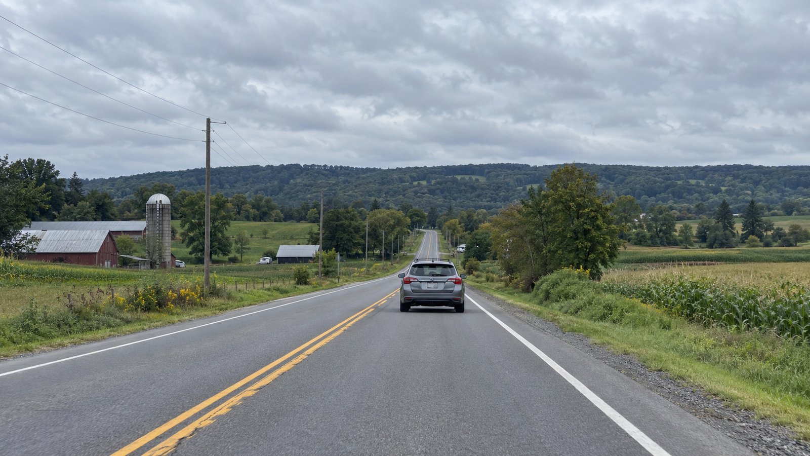 A gray station wagon travels in the right lane on a country road through low wooded farmland.