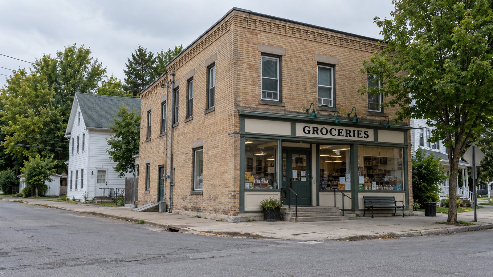 An everyday village street corner Two-story buff brick grocery storefront with entrance steps and a bench beside a quiet street corner