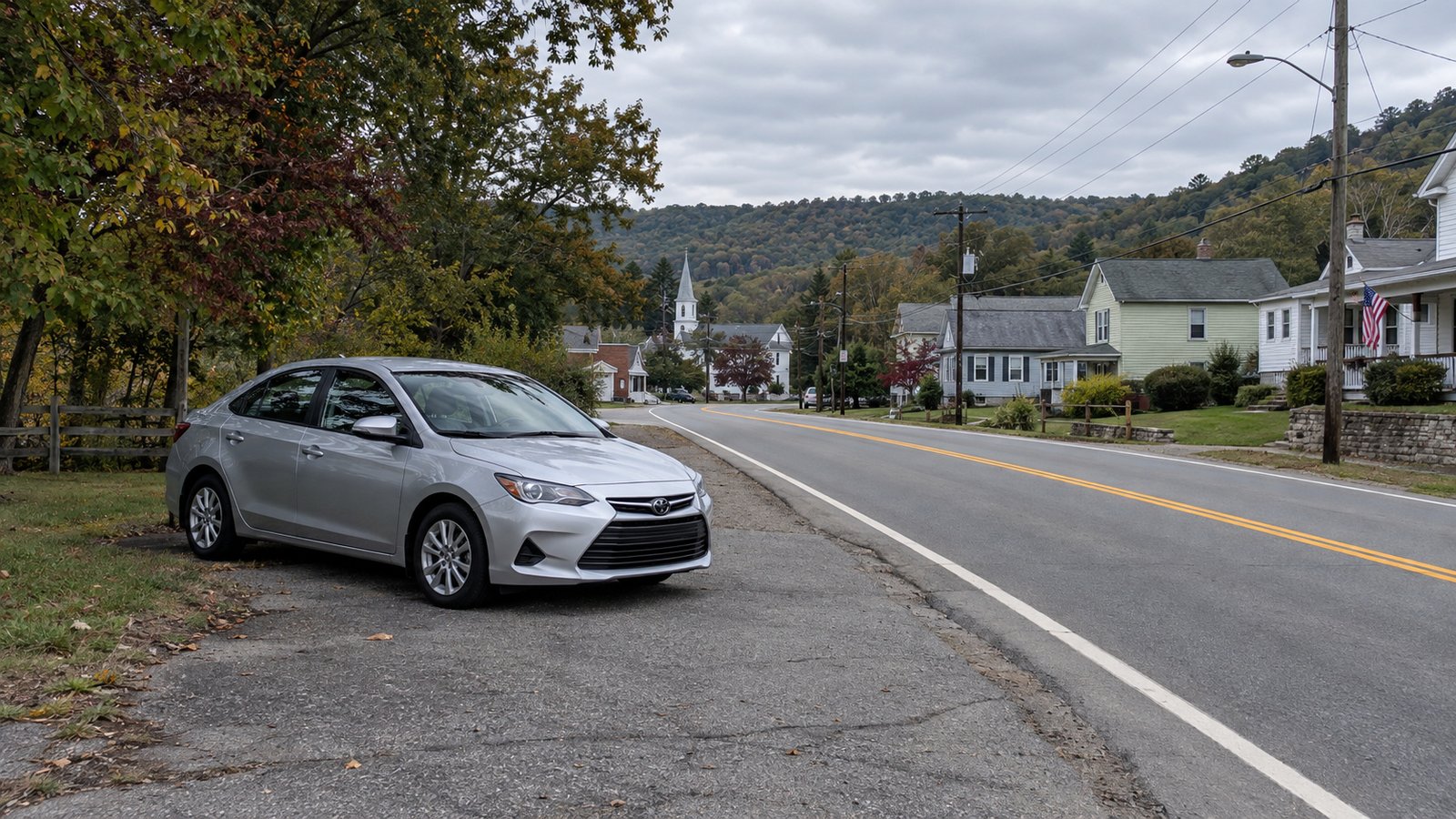 Trying a regular journey Silver car on a paved pull-off beside a two-lane road leading past village houses toward wooded hills
