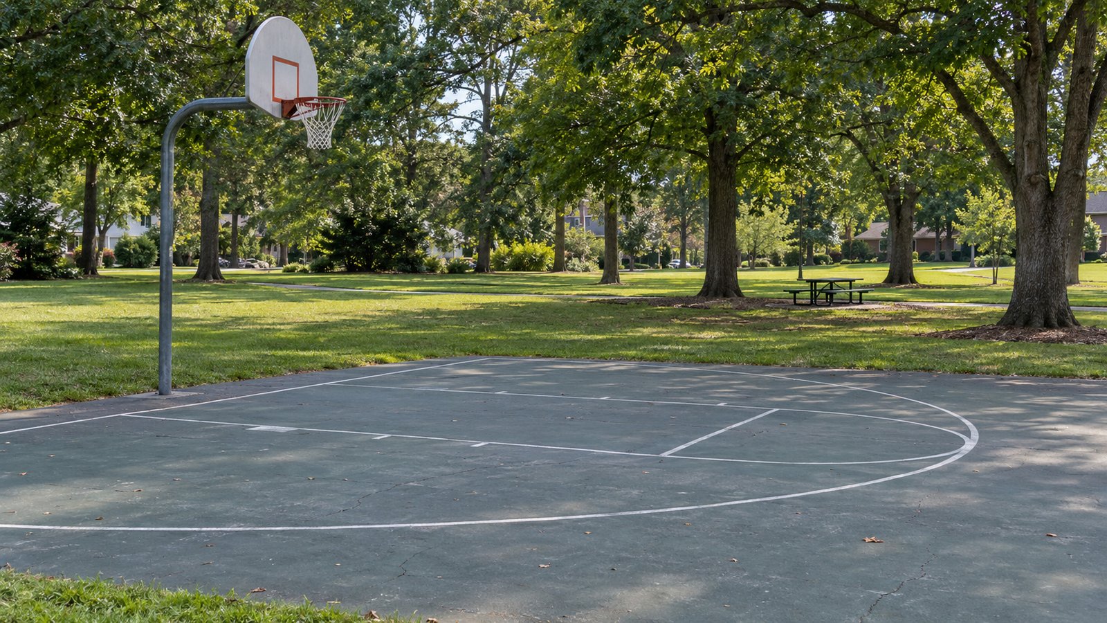 A neighborhood basketball court Empty outdoor basketball half-court beside grass, mature trees and a picnic table