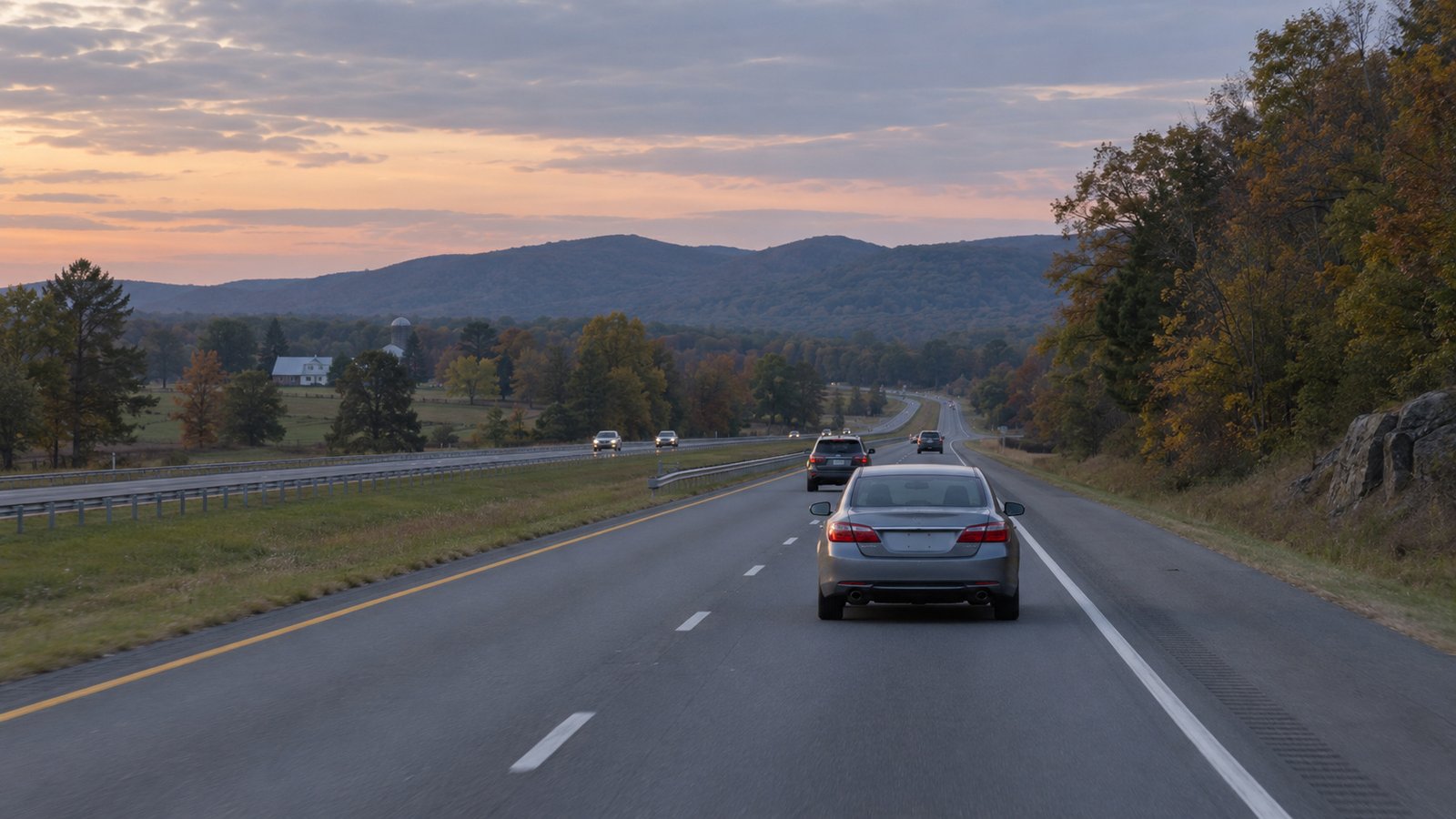 Gray sedan traveling away in the right-hand lane of a divided road beside low wooded hills.
