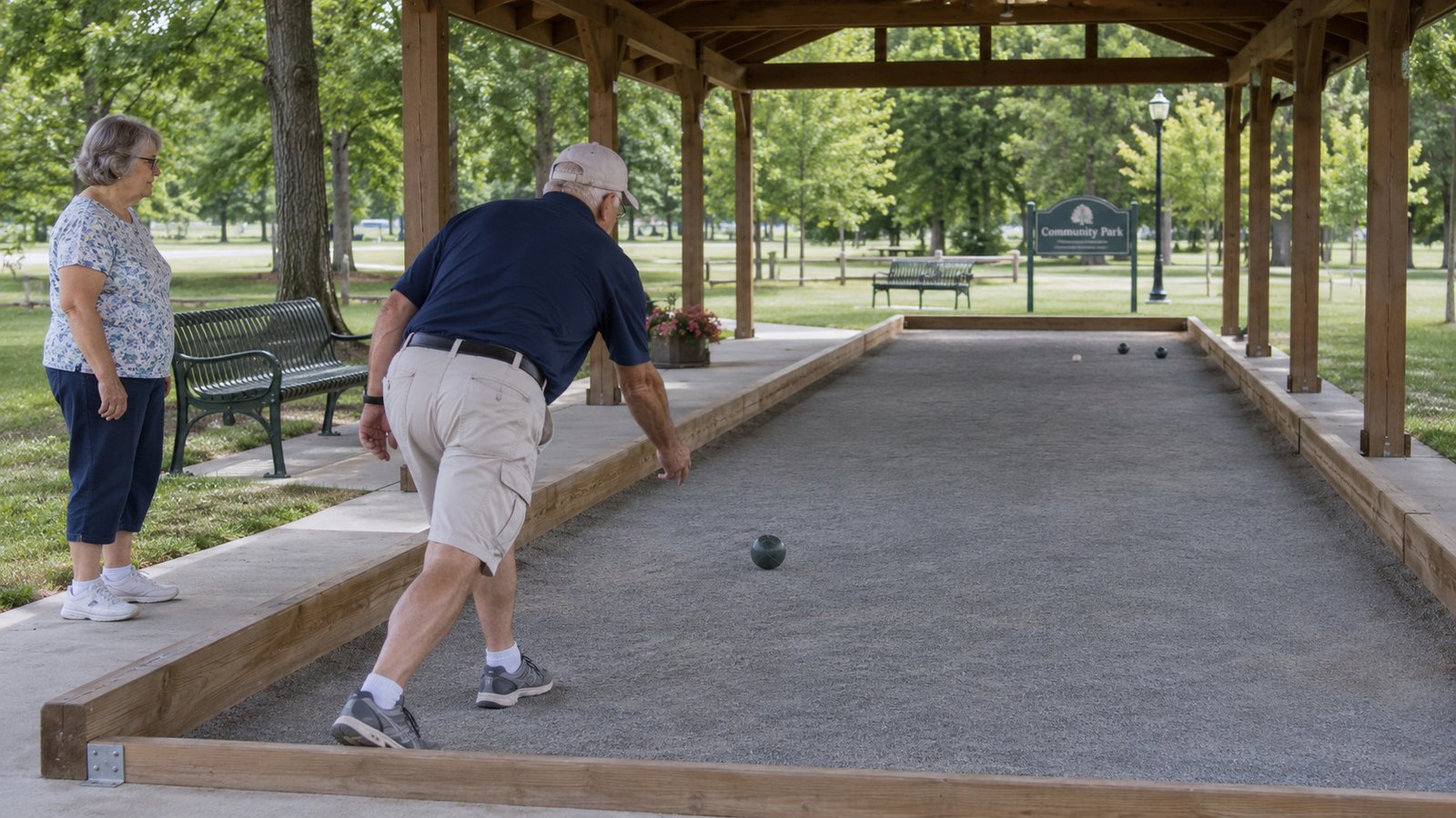Older man rolling a bocce ball while a woman watches beside a covered court.