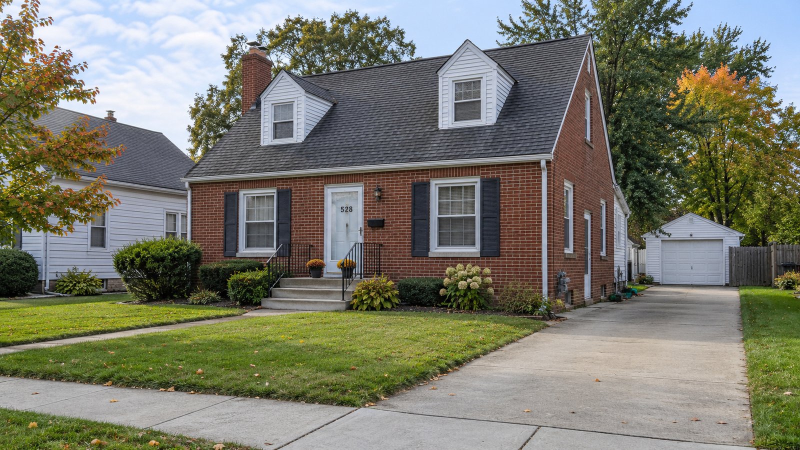 Brick Cape Cod house with two dormers, front steps, a side driveway and detached garage.