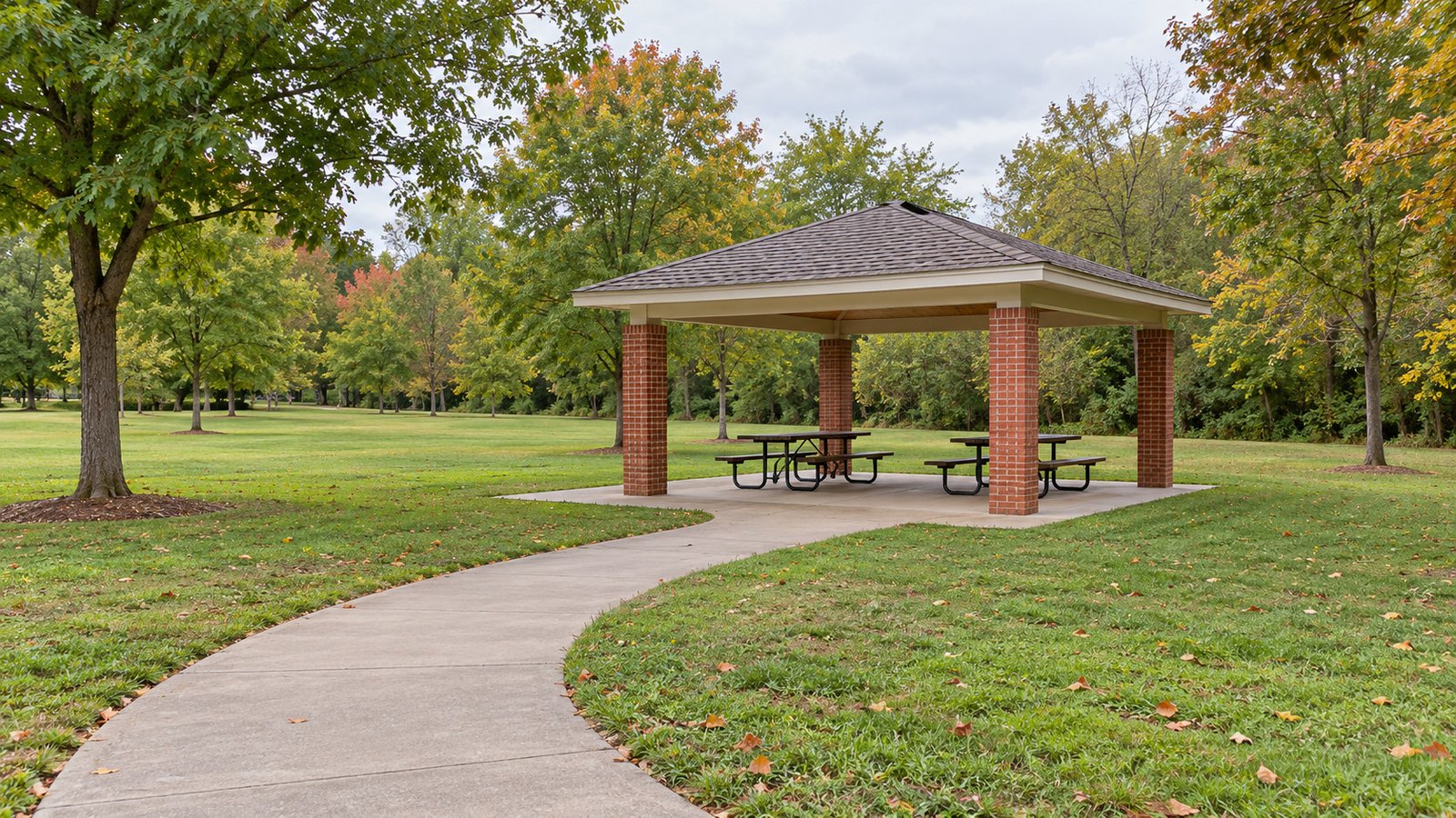 A paved park path joins a level picnic-pavilion slab with four brick posts and two tables.