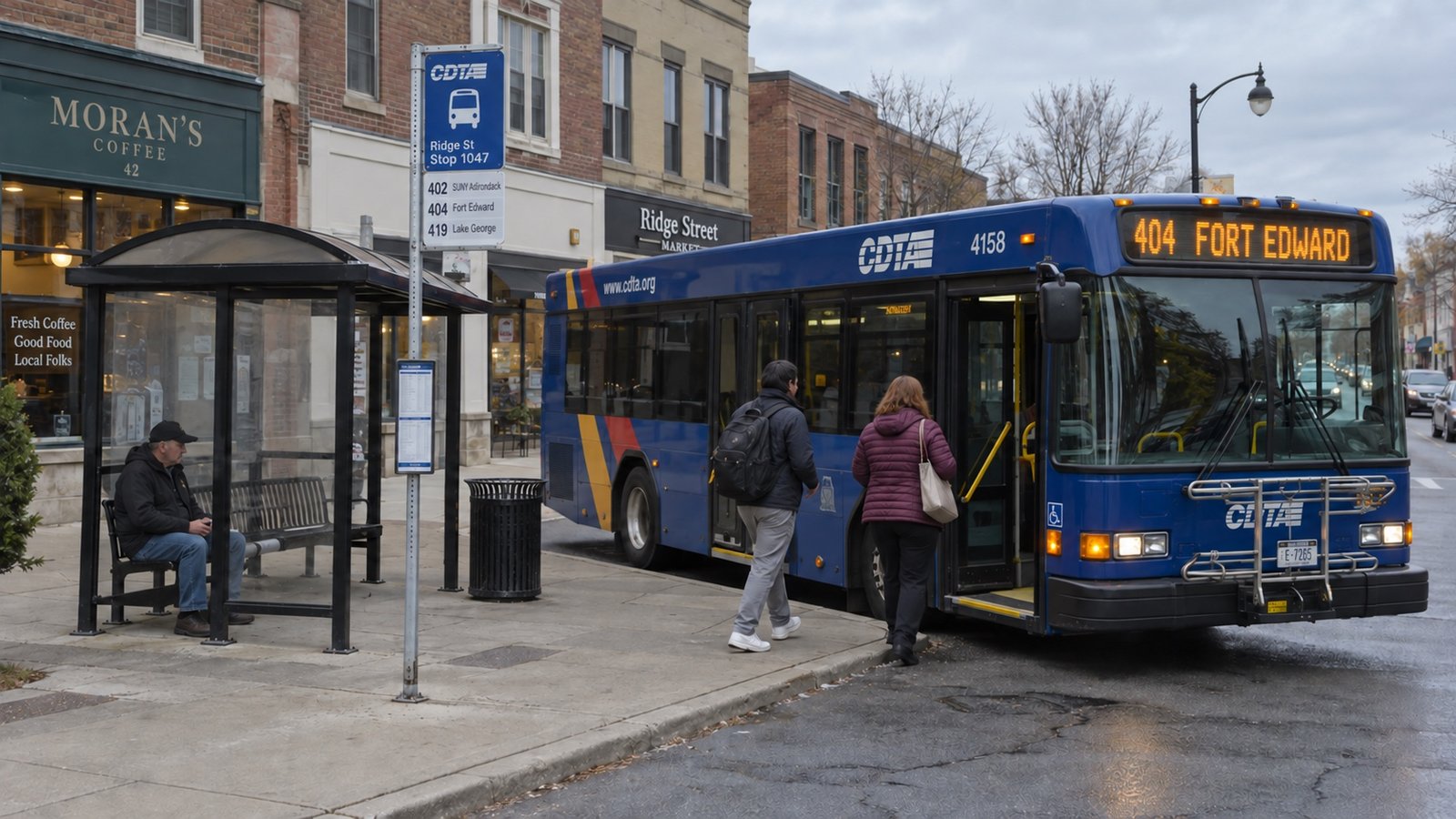 A city bus at a downtown stop beside low brick buildings and a terminal shelter.