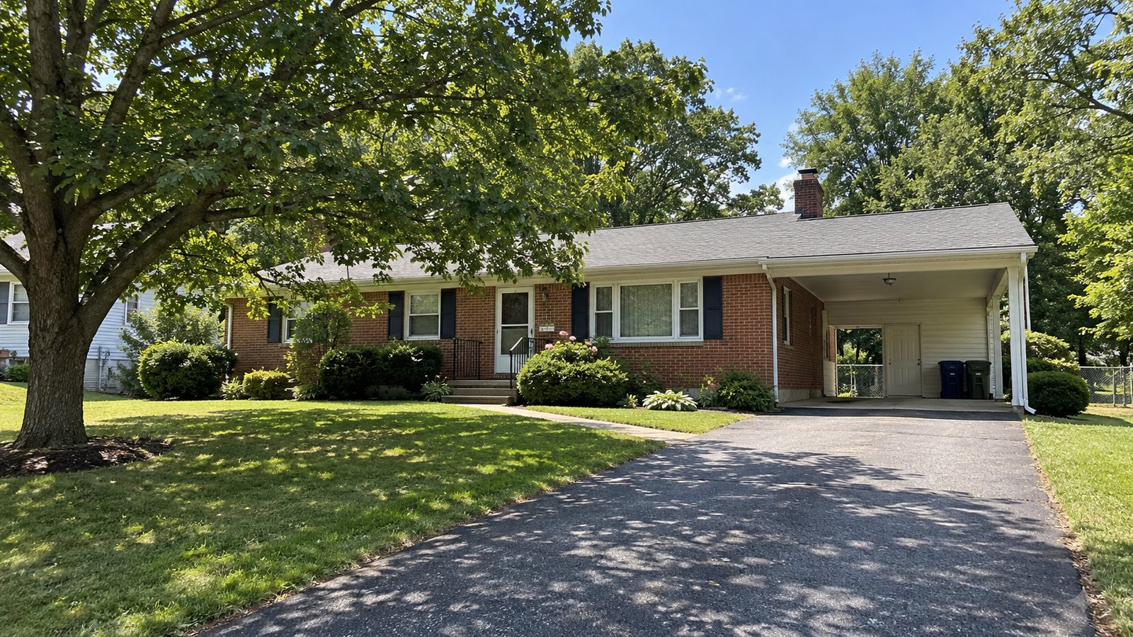 A complete one-story brick ranch house and attached carport beside a connected paved driveway under warm summer daylight.