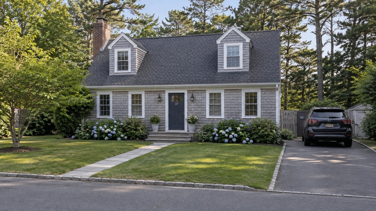 Planning a Hampton Bays home move A gray cedar-shingled house with white trim, flowering shrubs and a black SUV in the driveway