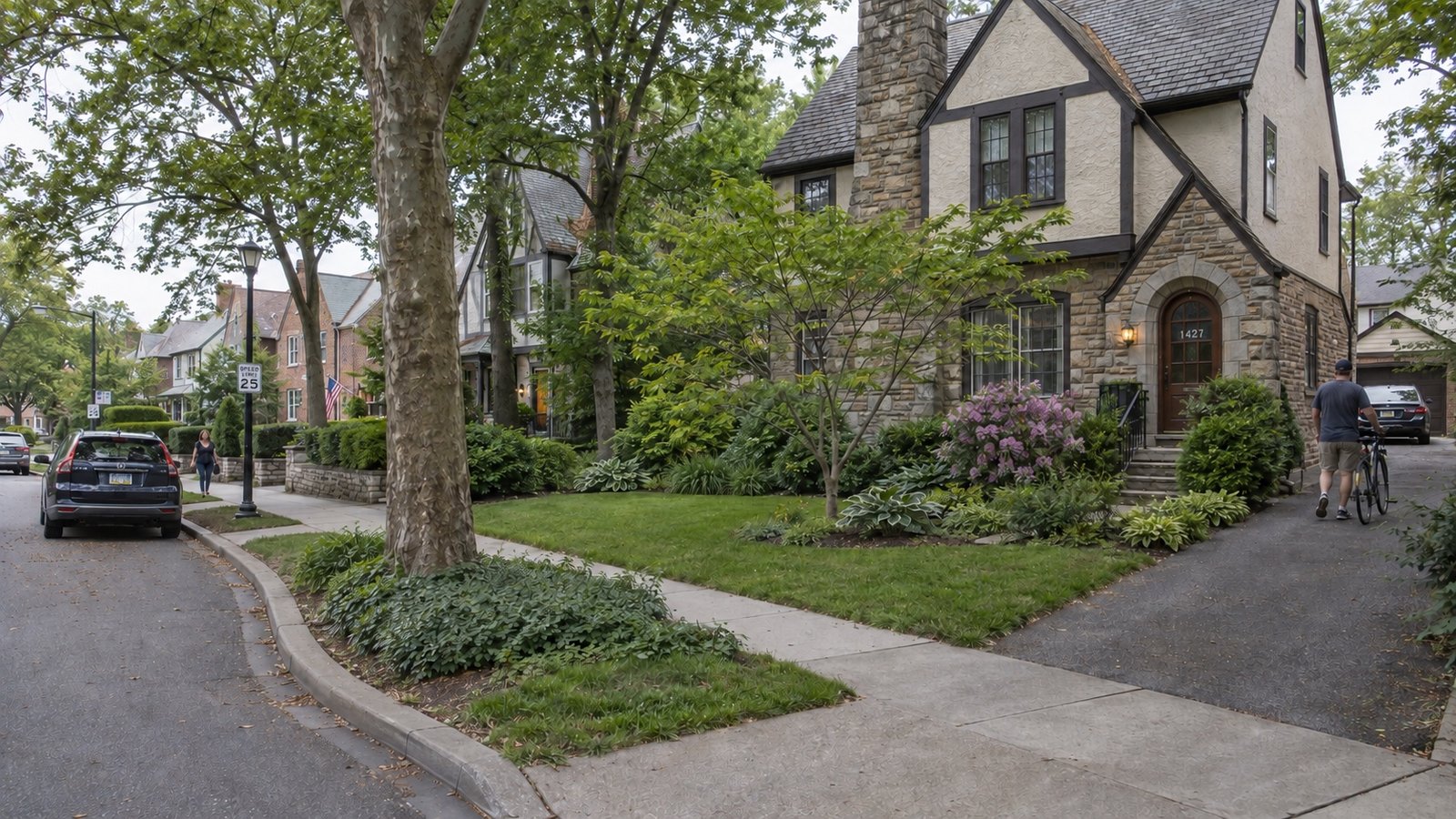 A stone-and-stucco house beside a landscaped curved residential street, with a resident wheeling a bicycle up the driveway.