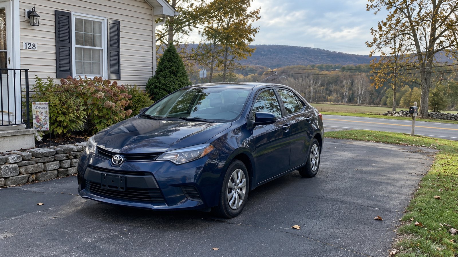 Planning daily journeys Blue compact sedan parked fully in a paved driveway beside a house with a road and wooded ridge beyond