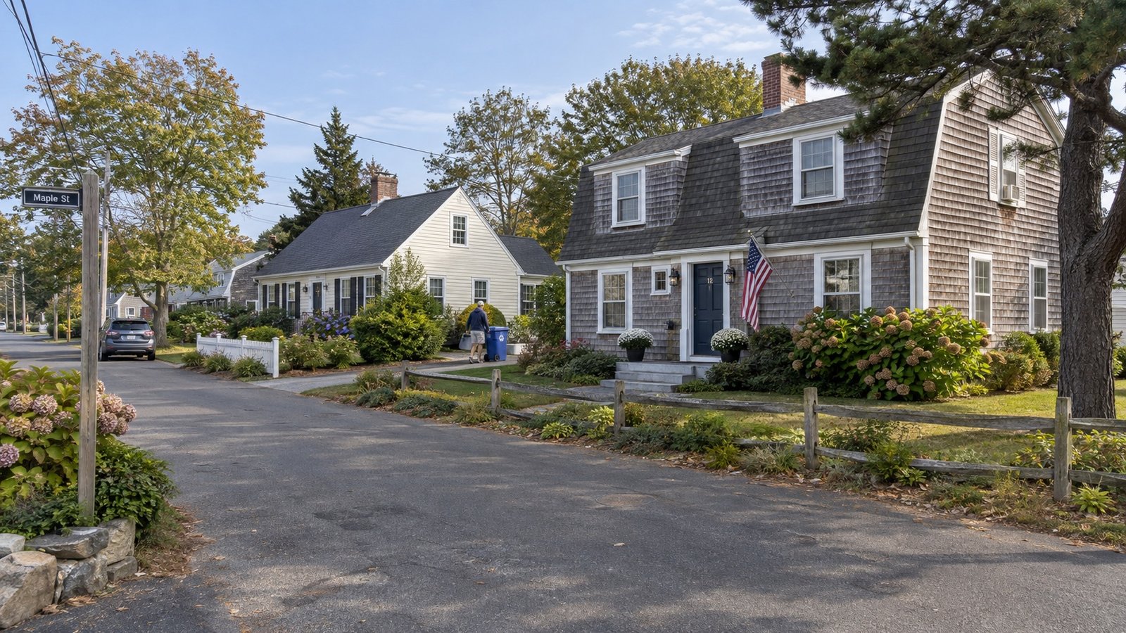 Shingled homes along a residential street Cedar-shingle homes beside a sloping Cape Cod neighborhood street, with a resident moving a recycling bin.