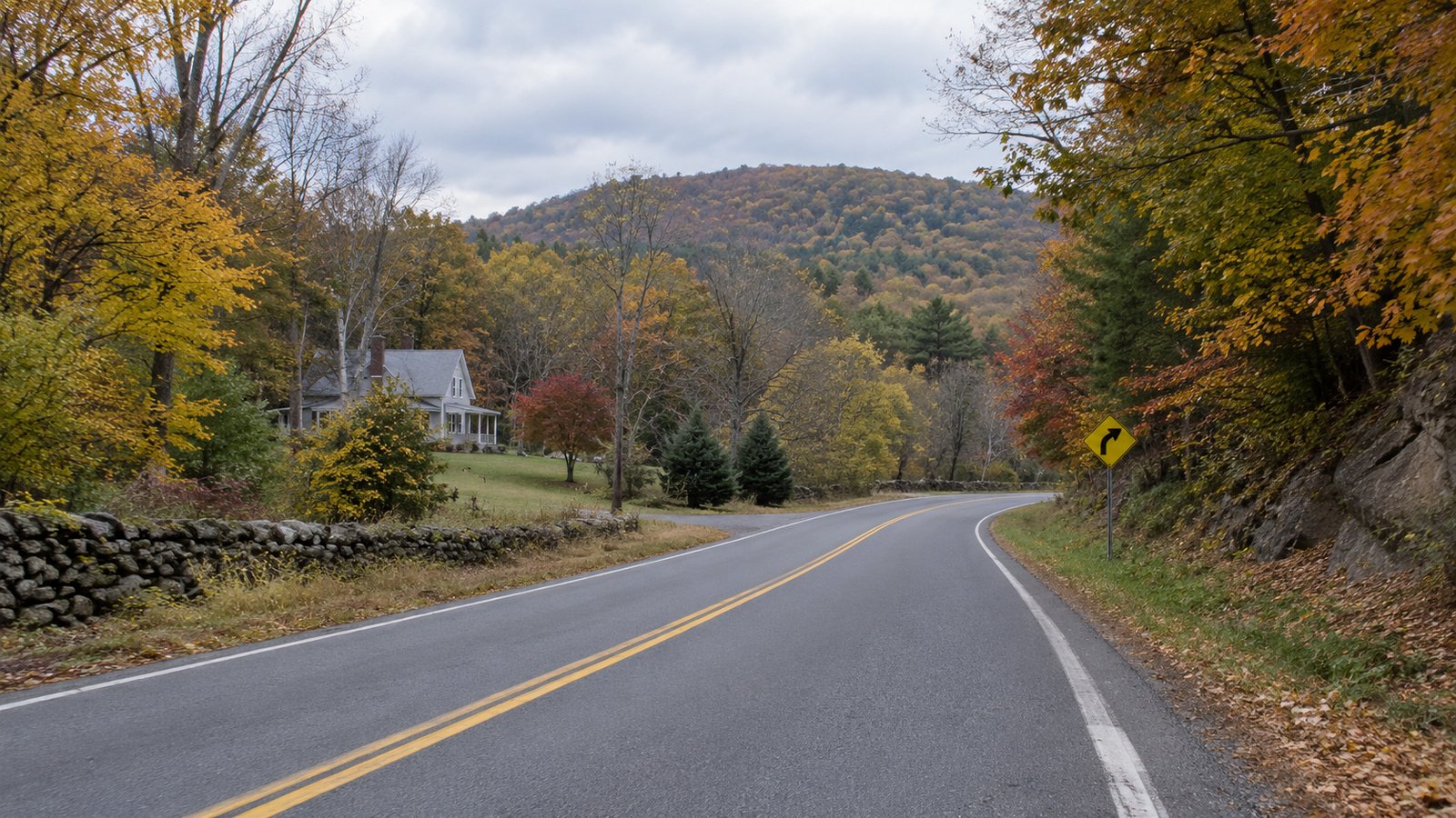 An empty curved road passes autumn trees, a roadside house and a low wooded ridge.