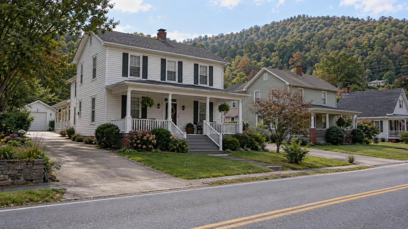 The home and its approach White two-story porch house with front steps, a paved driveway, yard and wooded hills behind.