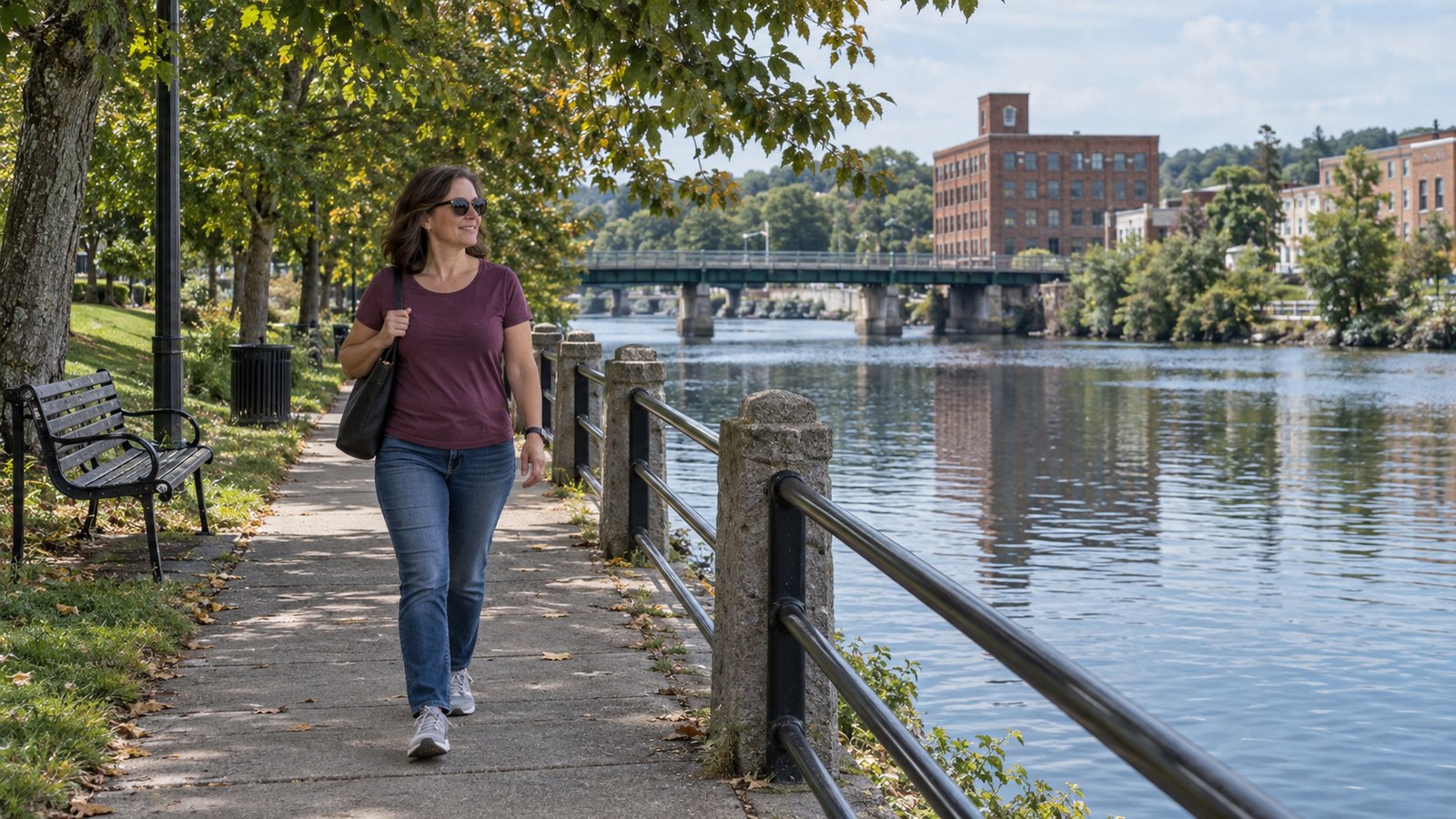 A woman walks a connected waterside path beside a railing and distant brick buildings.