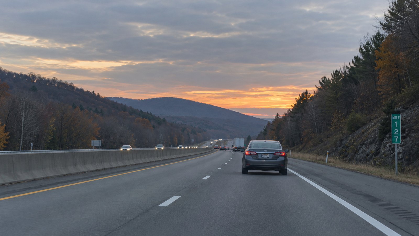 A whole compact sedan travels away on a divided highway through low wooded ridges at dawn.