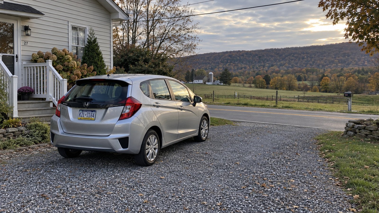 A silver hatchback parked on a gravel driveway beside a quiet valley road