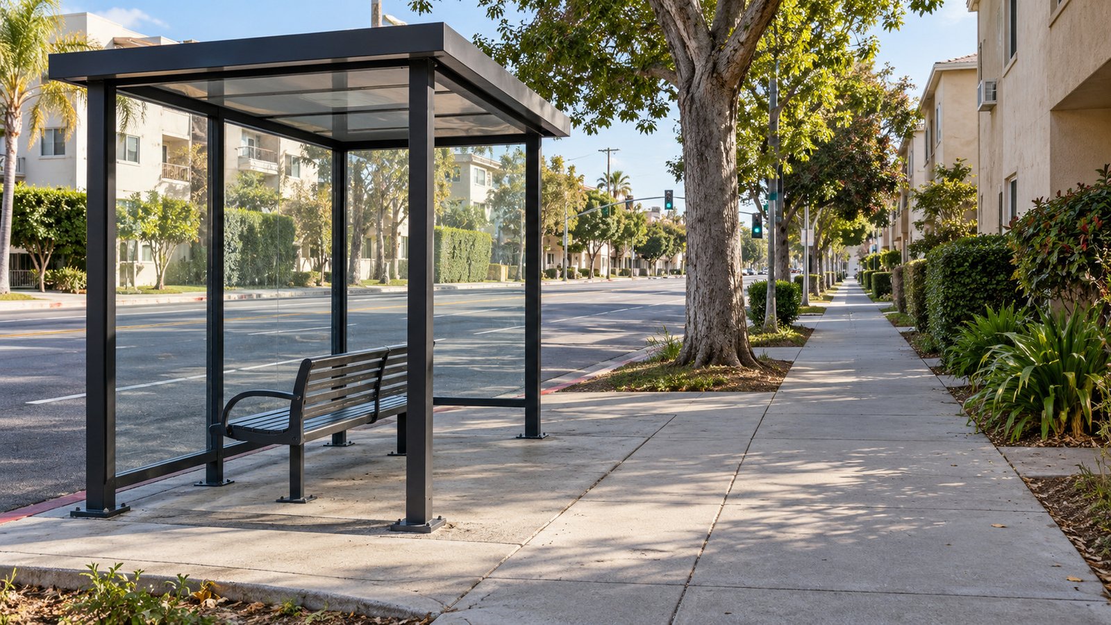 A complete glass bus shelter and supported bench in a separate sidewalk recess beside a continuous shaded walking route.