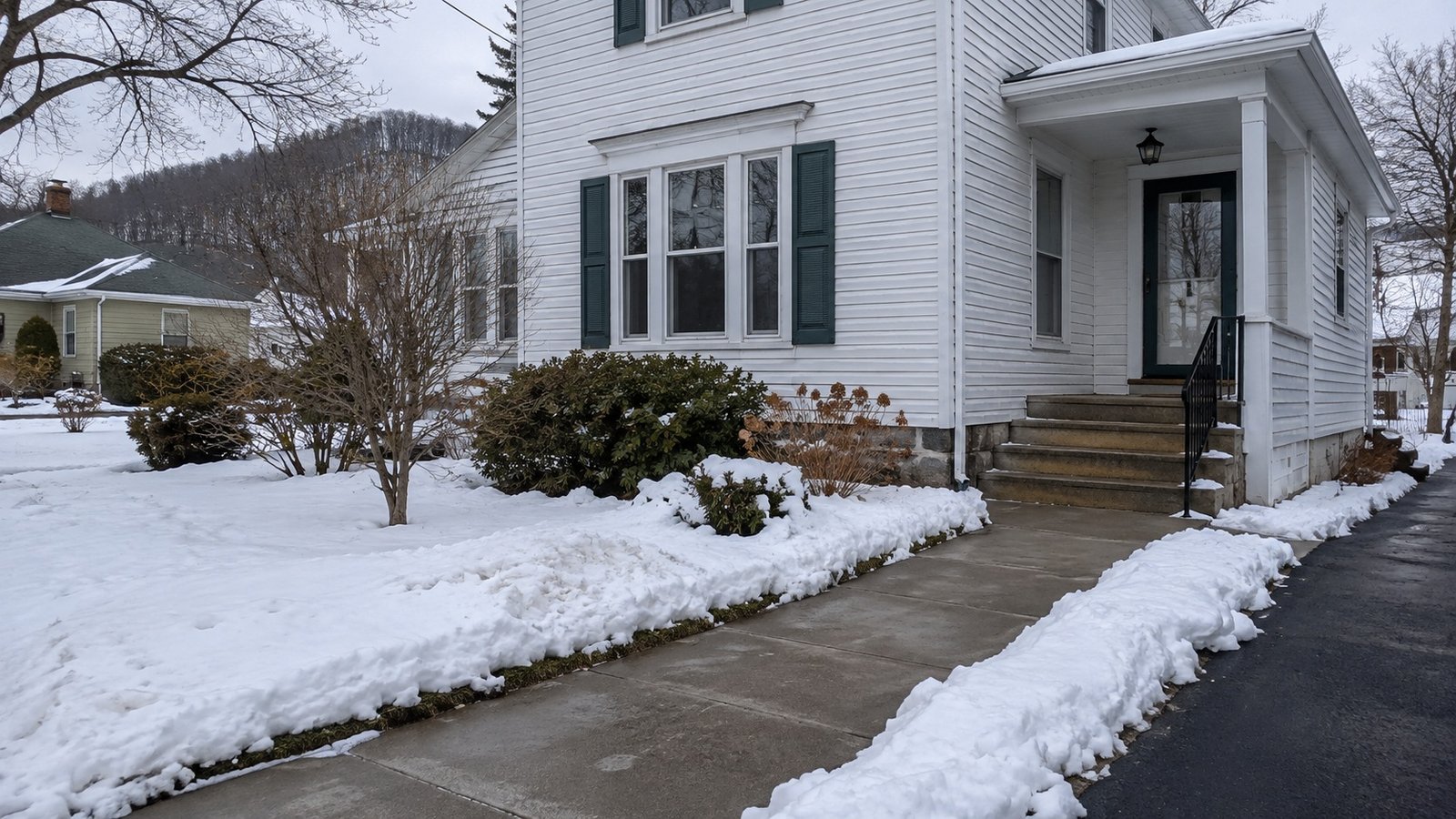 Cleared concrete walkway and porch steps at a white clapboard home, with snow along the path and an adjoining driveway
