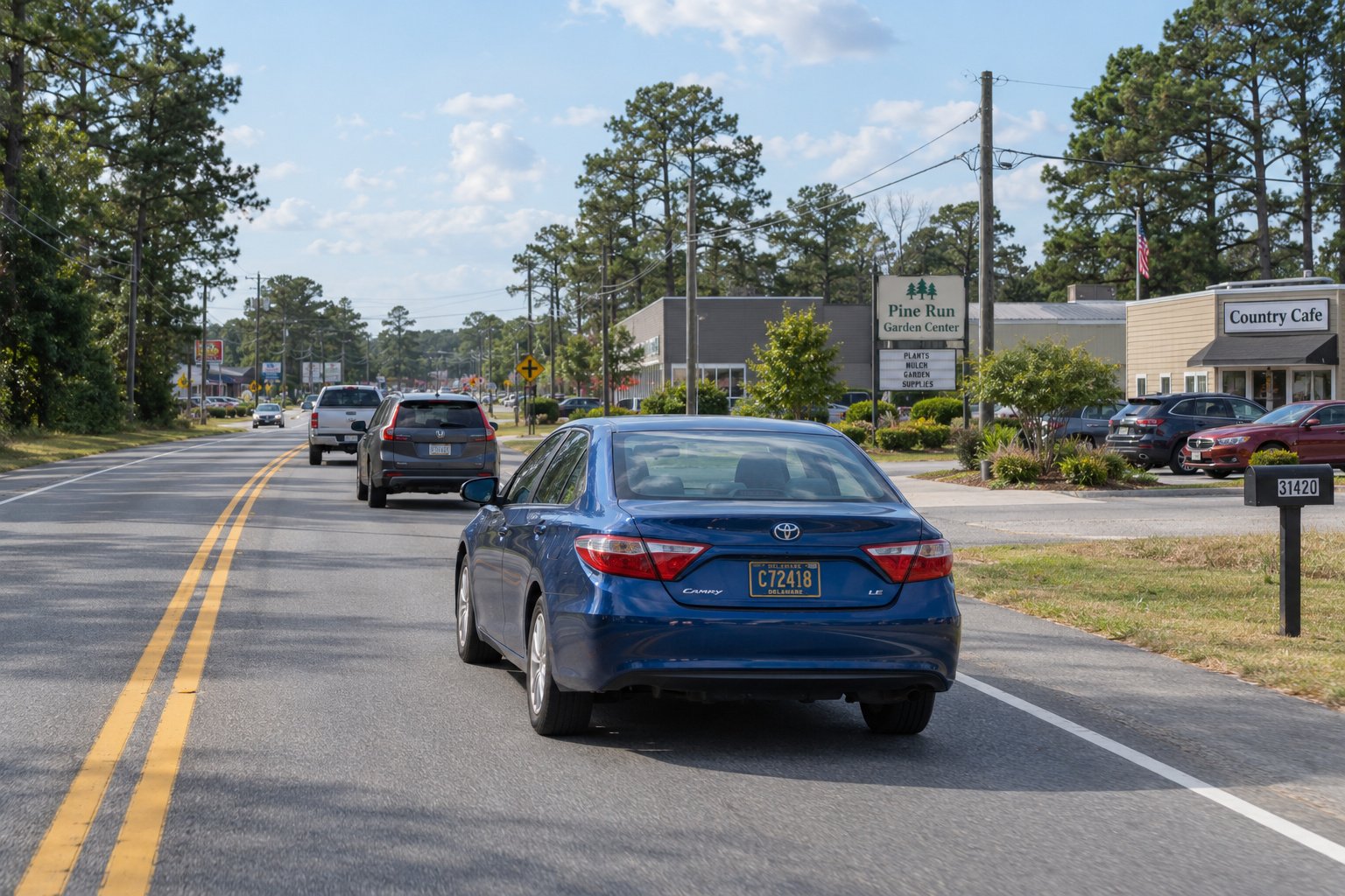 A complete blue sedan travels within the right lane of a level road beside ordinary shops and trees.