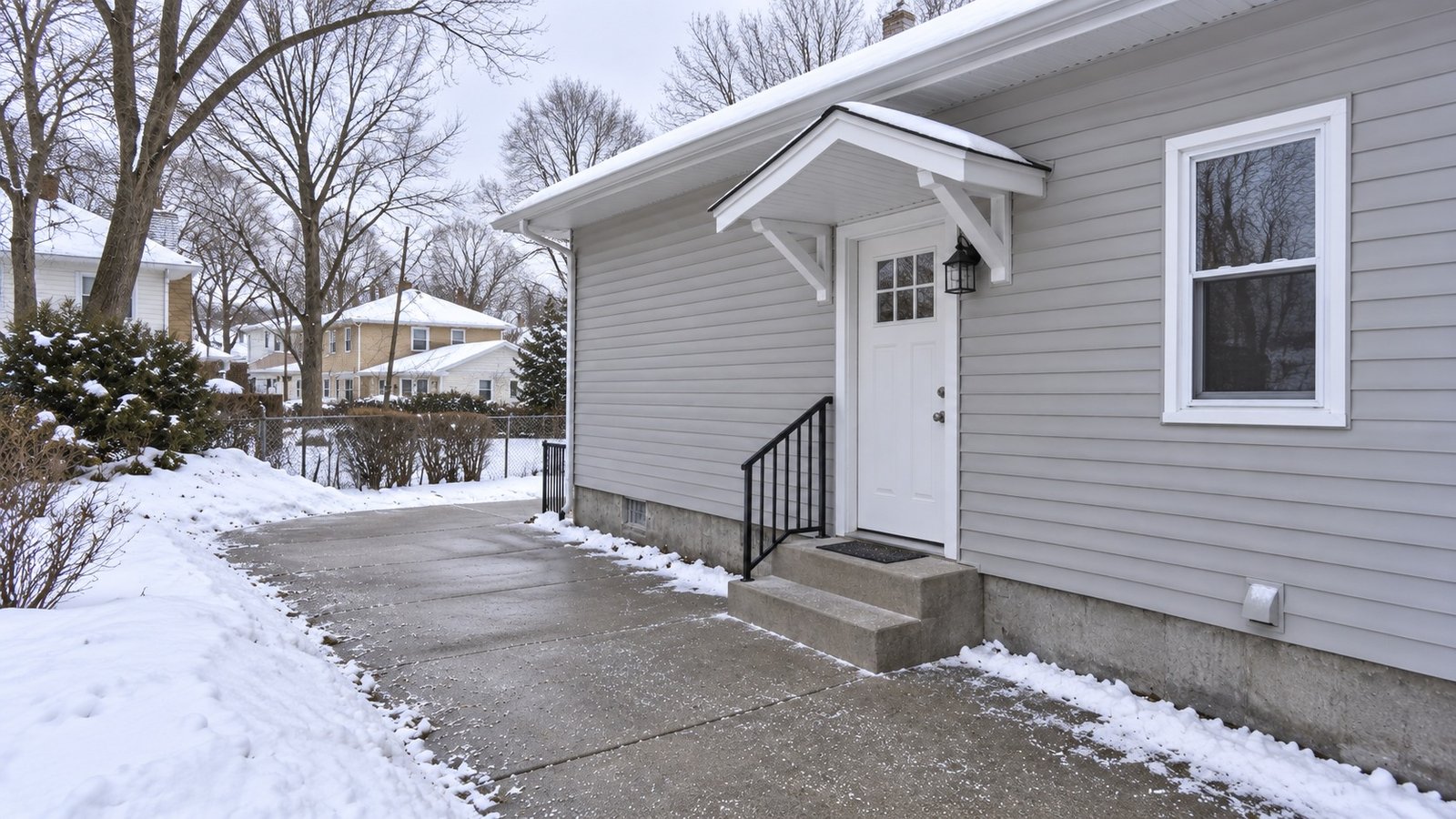 A white-sided home entry with a complete small overhang, two exposed concrete steps, a handrail and a path bordered by snowbanks.