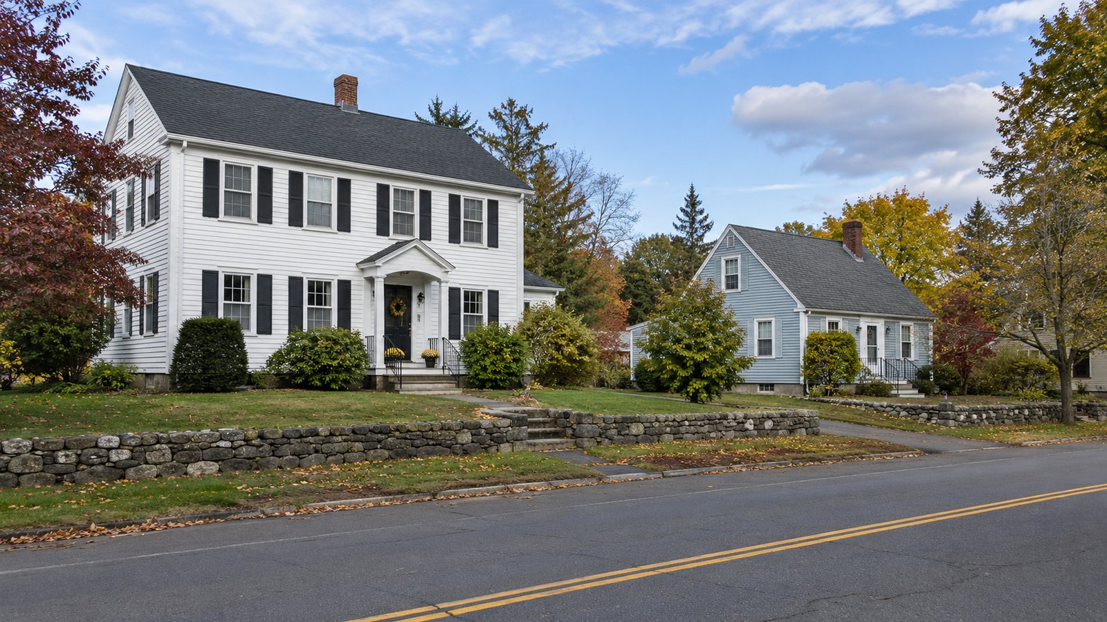 Two whole clapboard homes, one white colonial and one pale-blue cape, beside a quiet street and low stone walls.