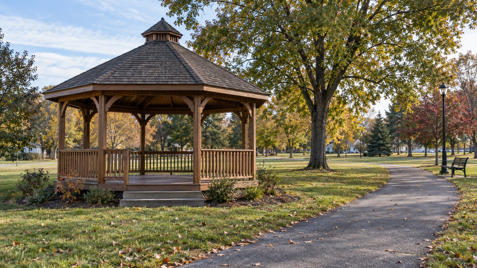 A whole timber gazebo beside a paved park path, lawn, autumn trees and a bench.