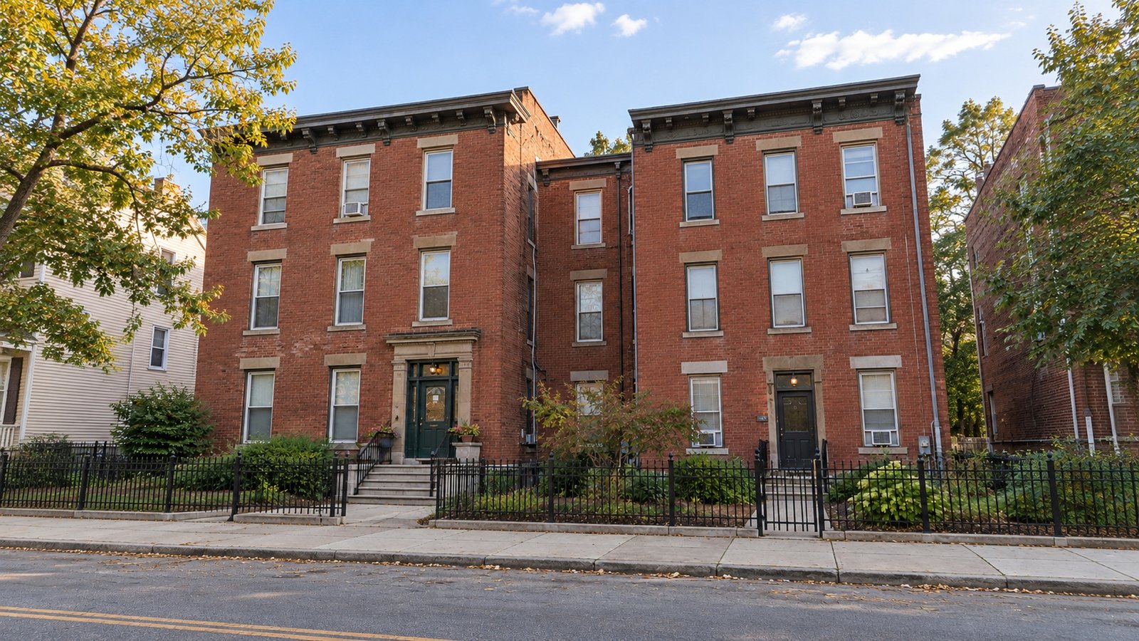 Show both the shared approach and the entrance when planning the carrying route. Two adjoining redbrick residential facades with separate doors, supported steps, iron gates and paths to the sidewalk.