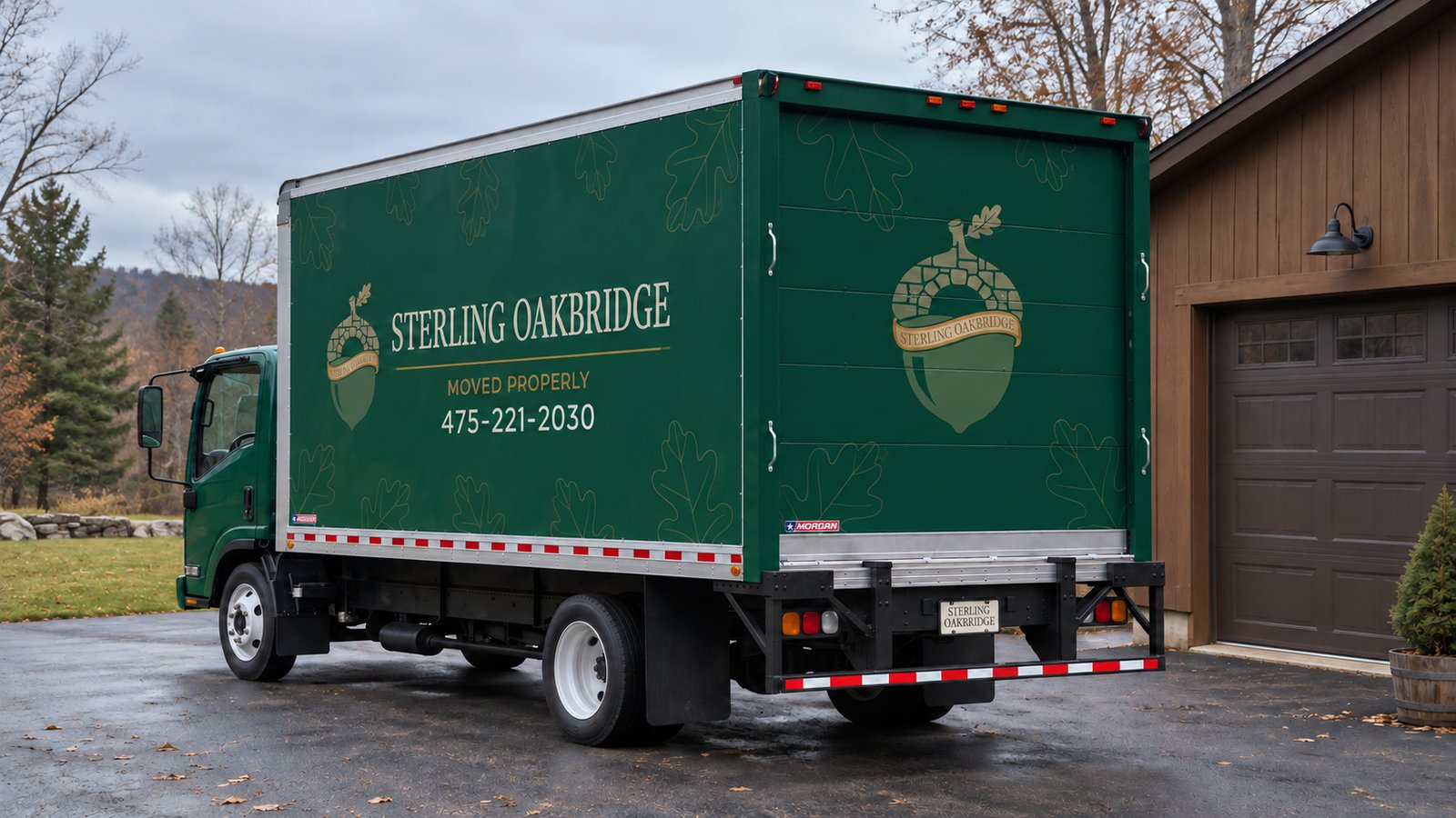 A position for the moving truck Dark green Sterling Oakbridge box truck viewed from the rear and left side on a private paved drive beside a wooden garage