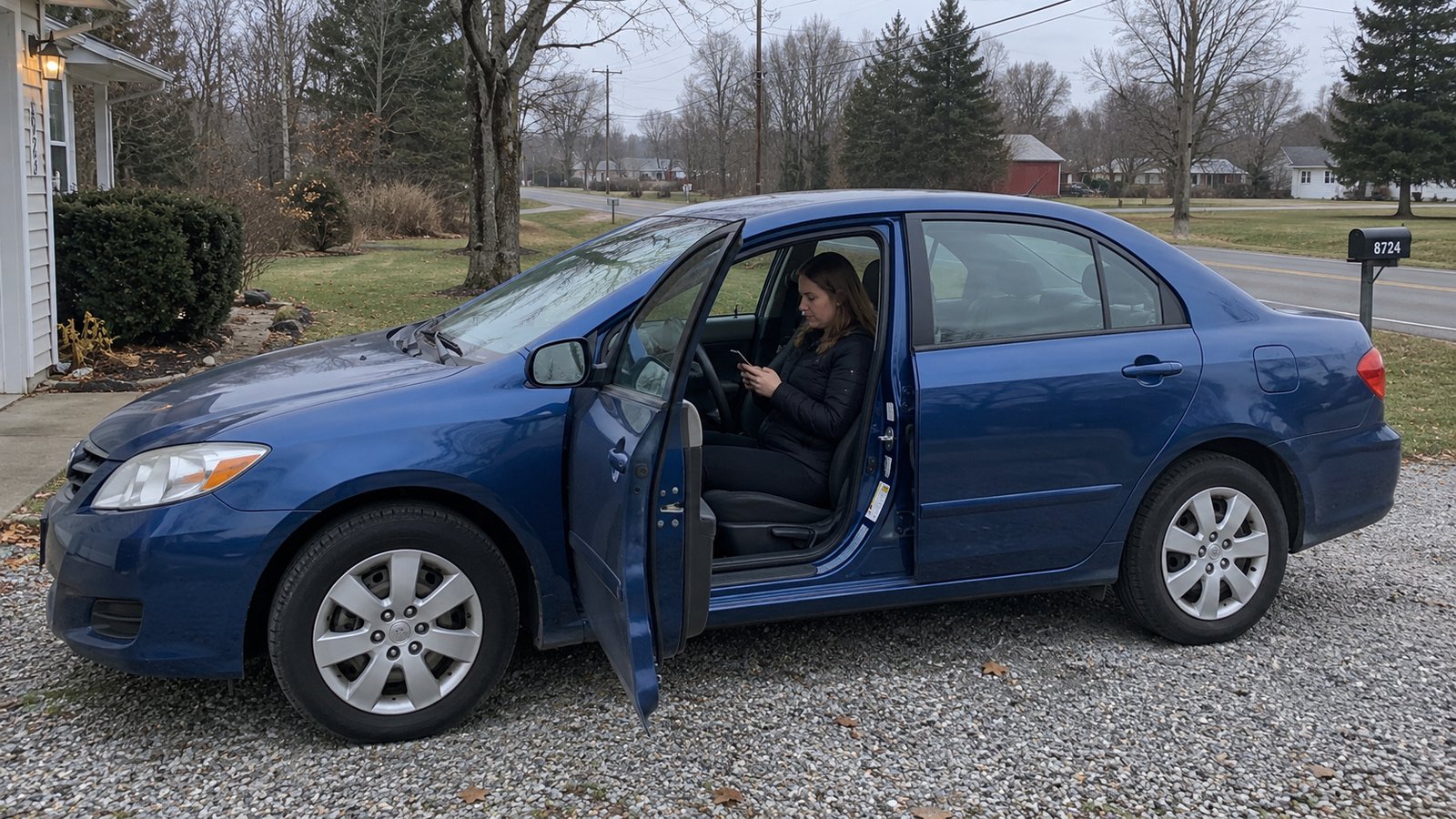 Woman consulting a phone in the driver seat of a stationary blue sedan with its door open on a gravel driveway.