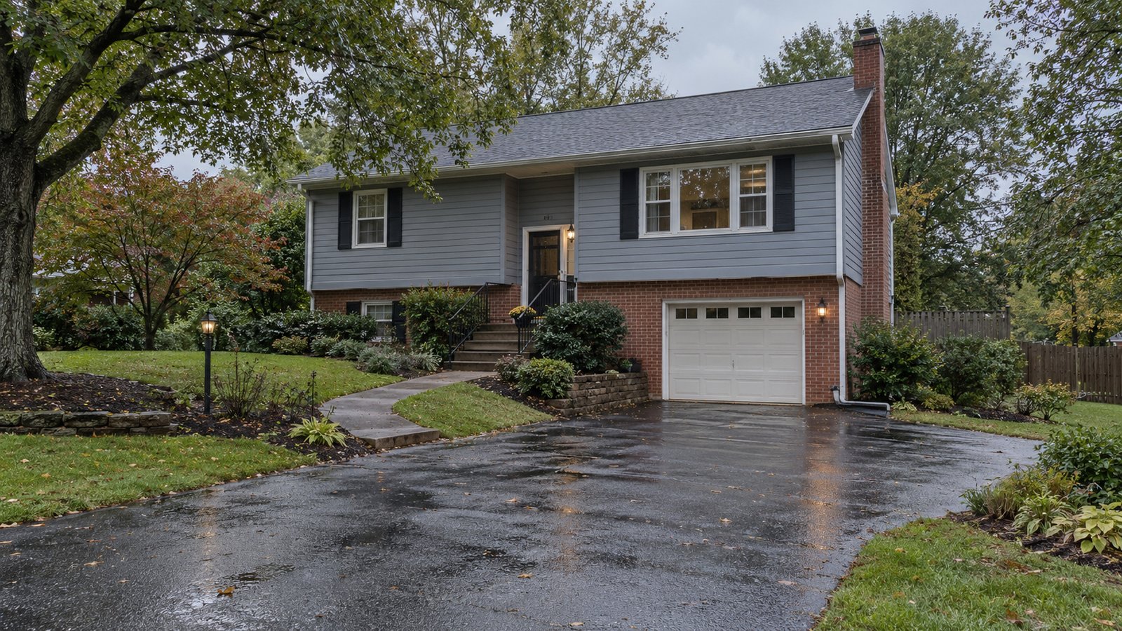 Review the approach after rain A complete gray-blue and brick split-level home beside a wet driveway and front walk after rain.