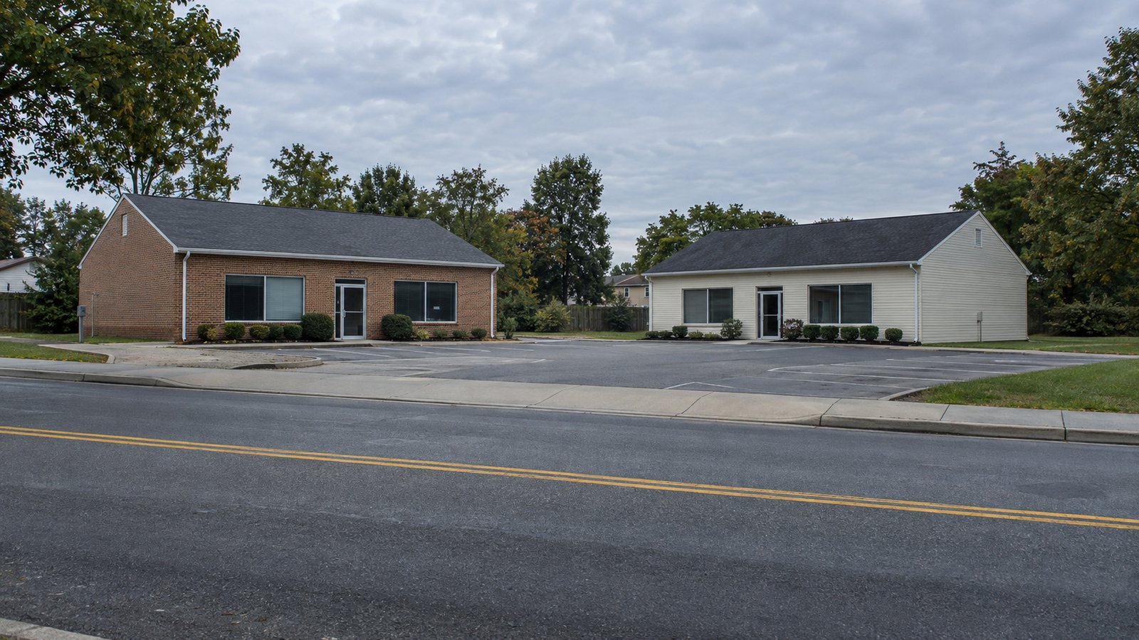 Two low buildings beside a paved forecourt and a sidewalk on an ordinary suburban street.