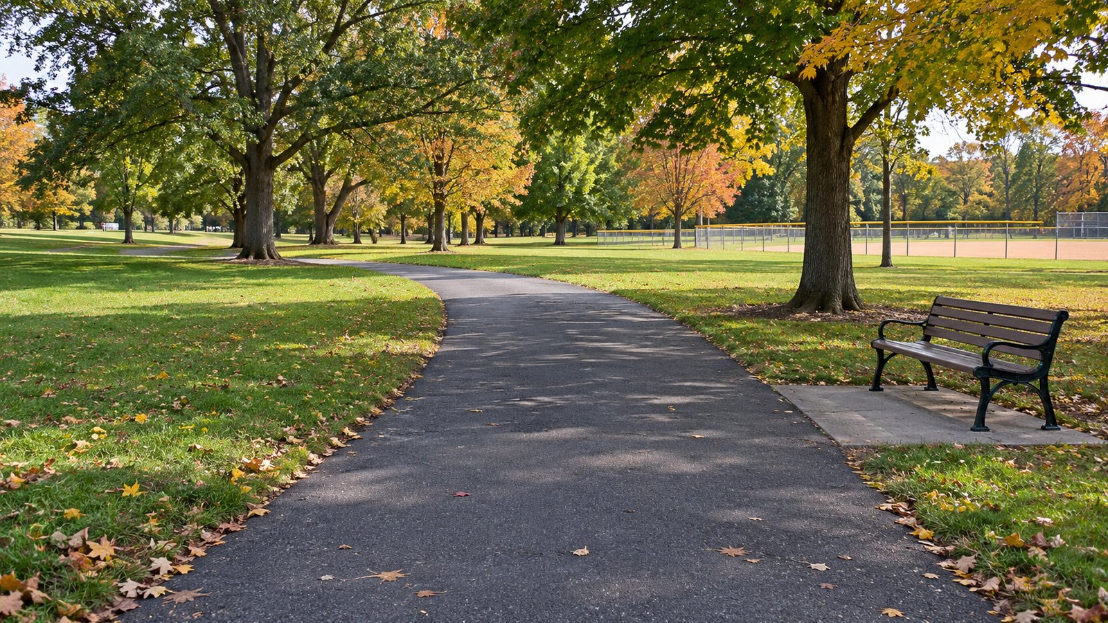 A curved paved park path, with a supported bench in a separate recess and a ballfield beyond the lawn.