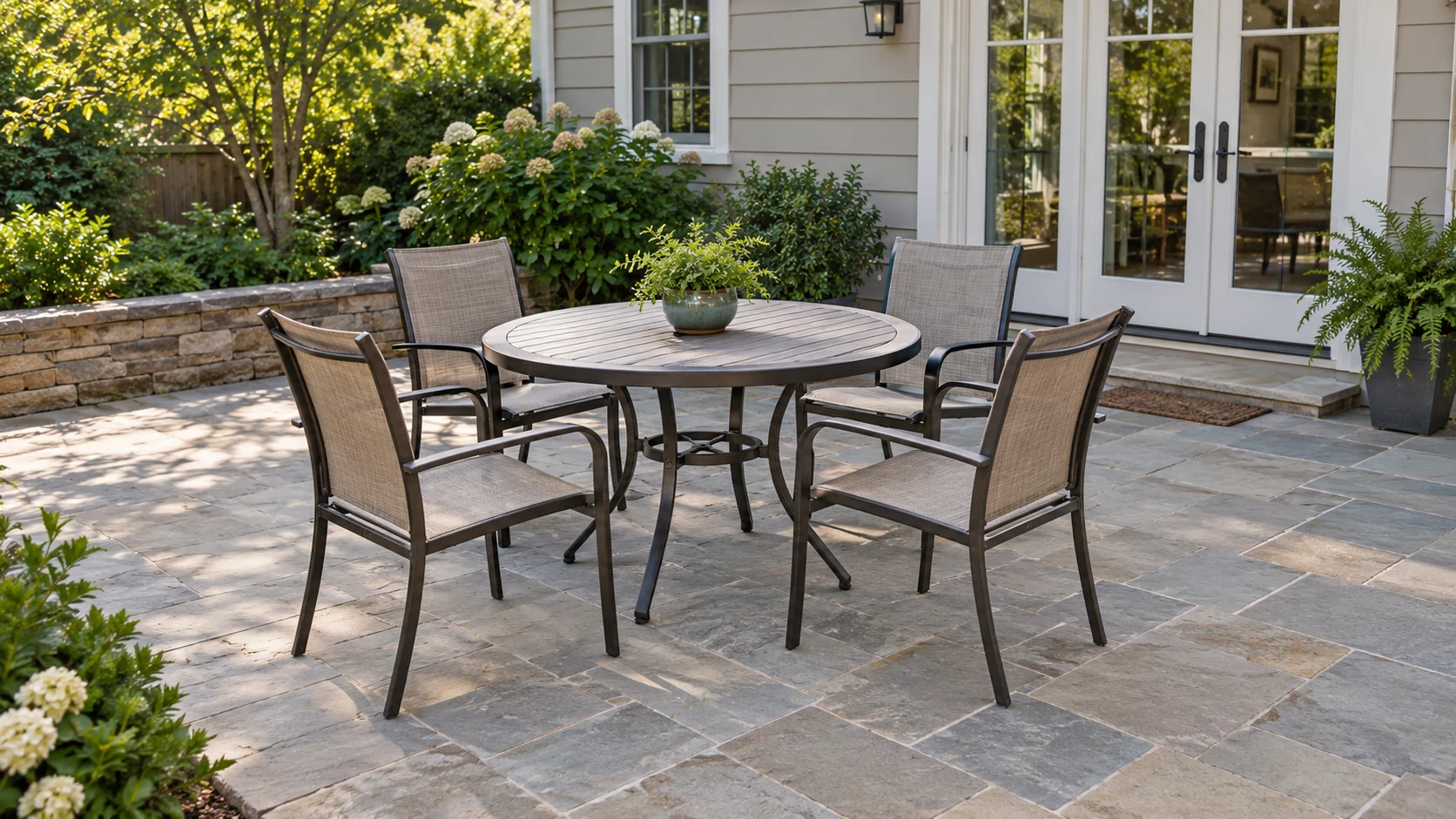 A round outdoor table and four chairs stand on a furnished patio beside a house and planted garden edge.