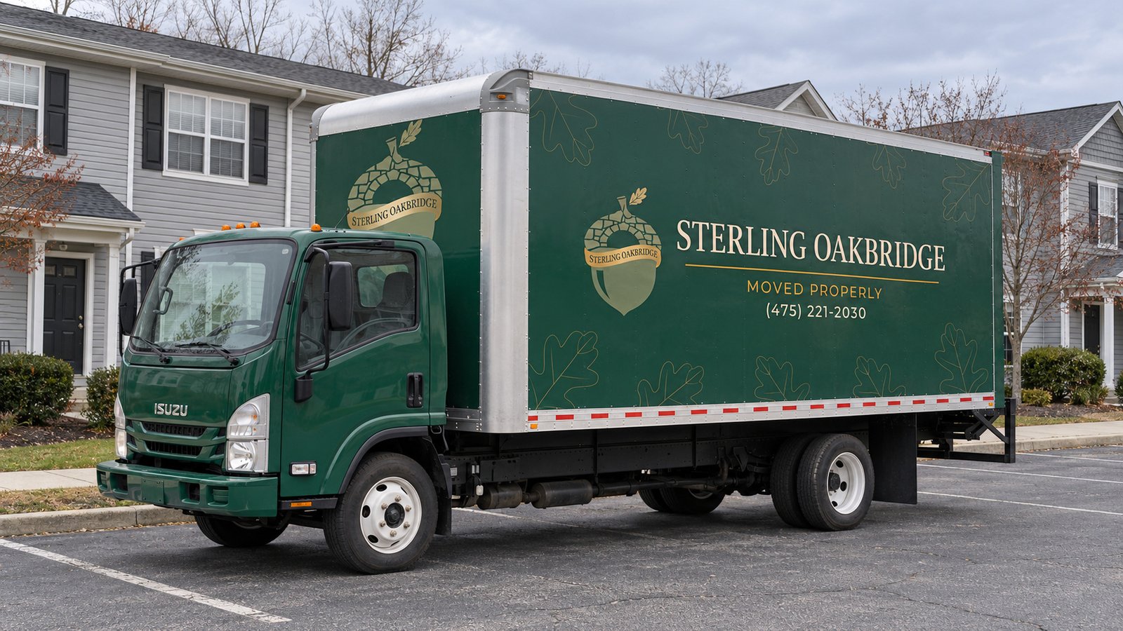 Vehicle position beside shared residential access Green Sterling Oakbridge Isuzu box truck on a paved parking area beside a row of gray-sided homes and a sidewalk